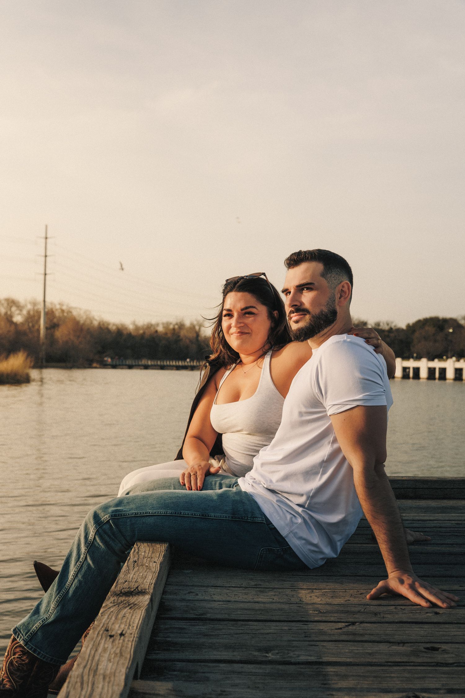 Couple sitting together on wooden dock by lake during sunset in casual white outfits.