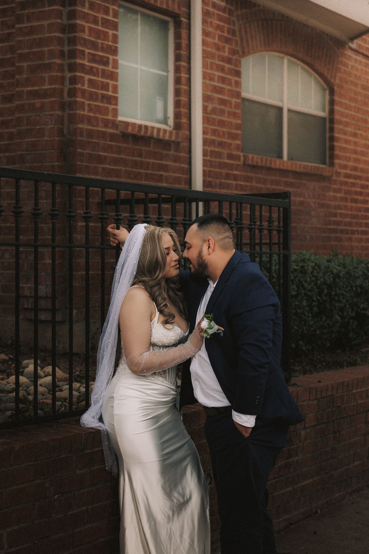A romantic wedding moment captured against a brick wall with black iron railing, featuring a bride in white gown and flowing veil.