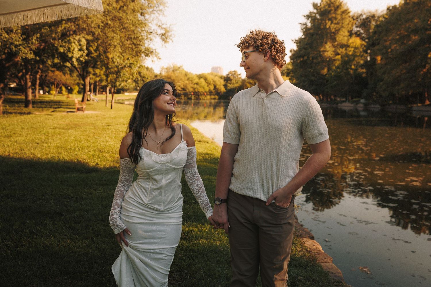 A couple walks hand in hand by a pond on an autumn day in a park setting.