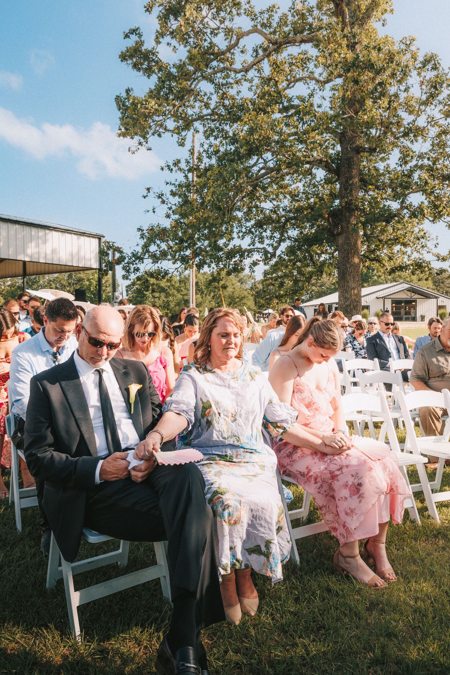 Wedding guests seated outdoors at a summer ceremony with white chairs under a shady tree.