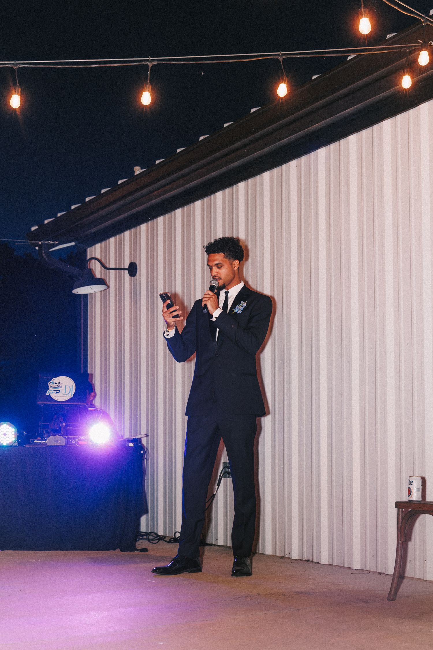 A groomsman in a navy suit delivers a speech at an evening wedding reception under string lights.