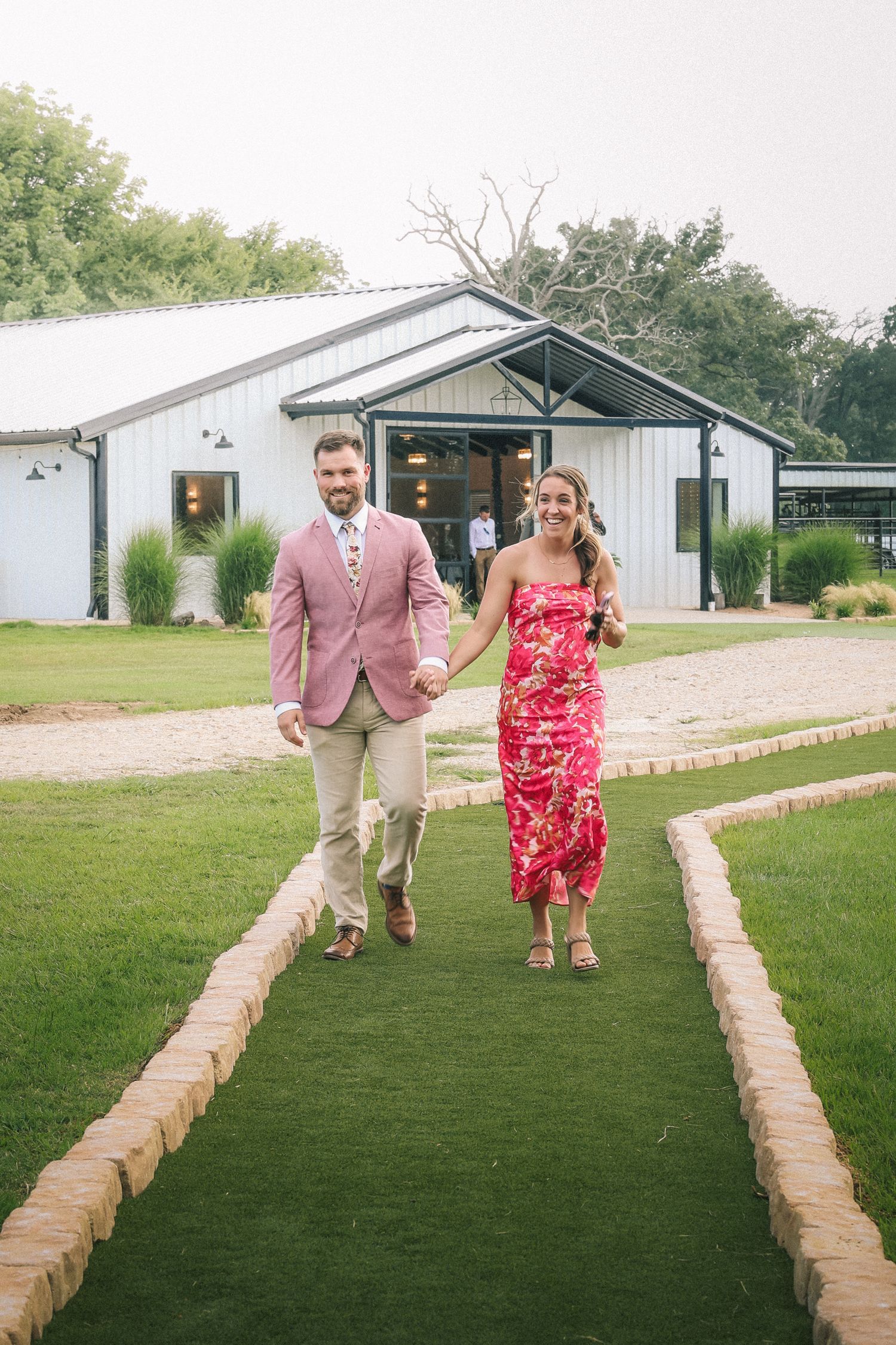 Wedding party walks down a stone-lined path between white buildings on a sunny day.