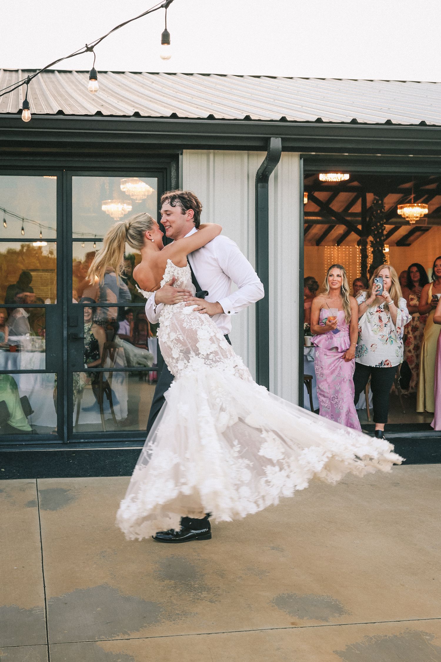 Newlyweds share their first dance while wedding guests watch through glass windows in the background.