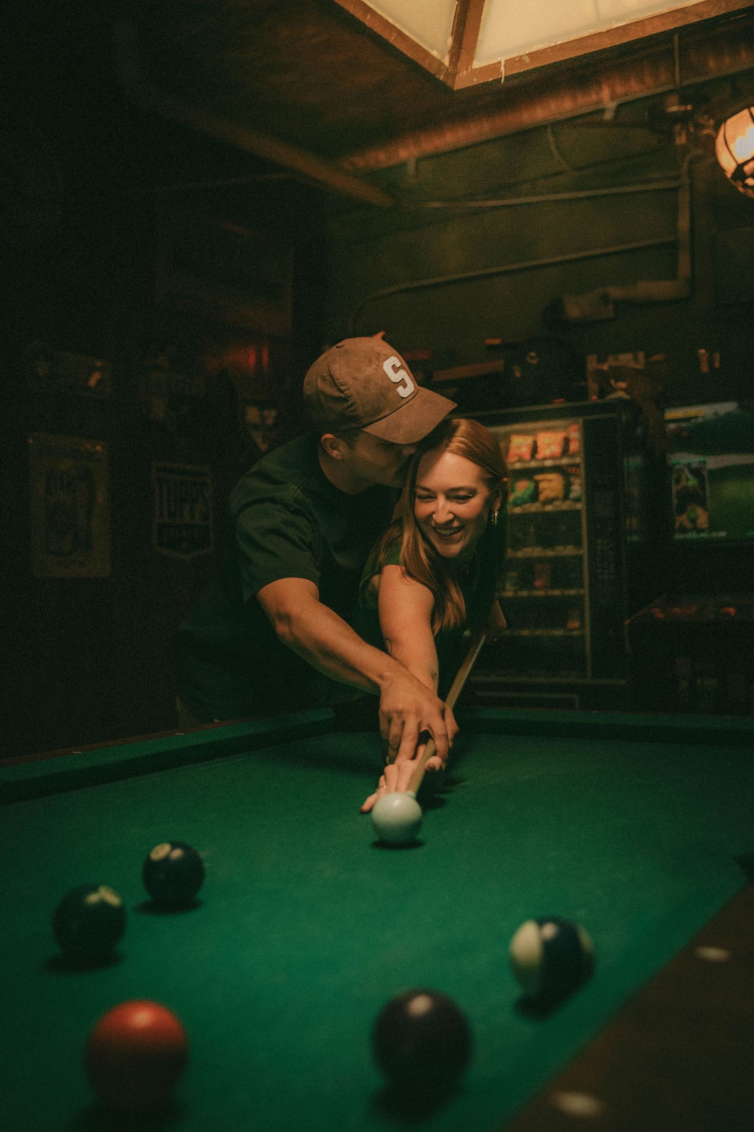 A person wearing a baseball cap lines up a shot at a pool table with green felt under warm lighting.