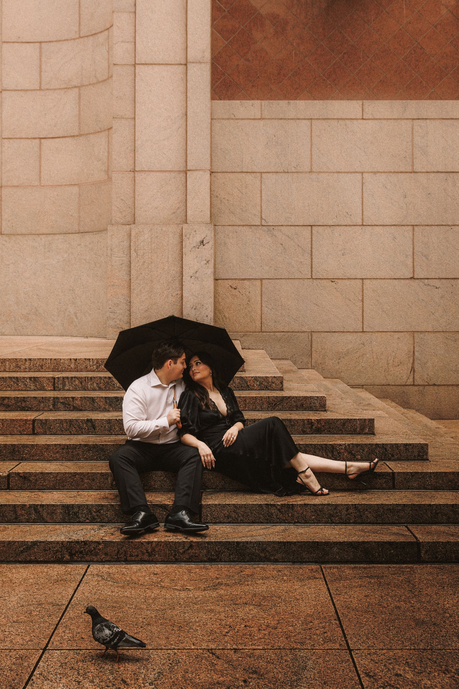 Romantic couple sharing a kiss under black umbrella on marble steps with a pigeon nearby.
