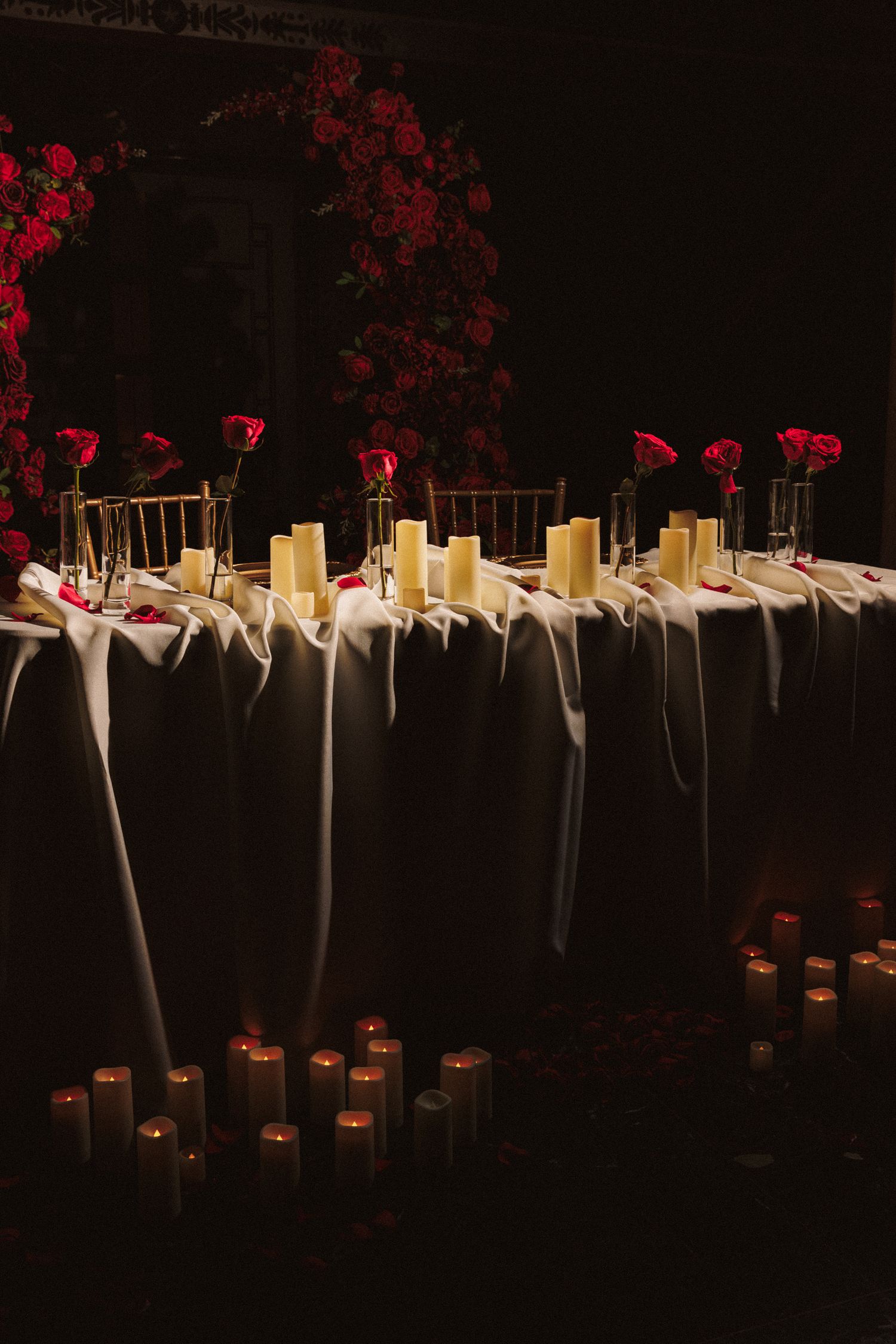 Elegant wedding reception table with white draping, pillar candles, and red roses in dim romantic lighting.