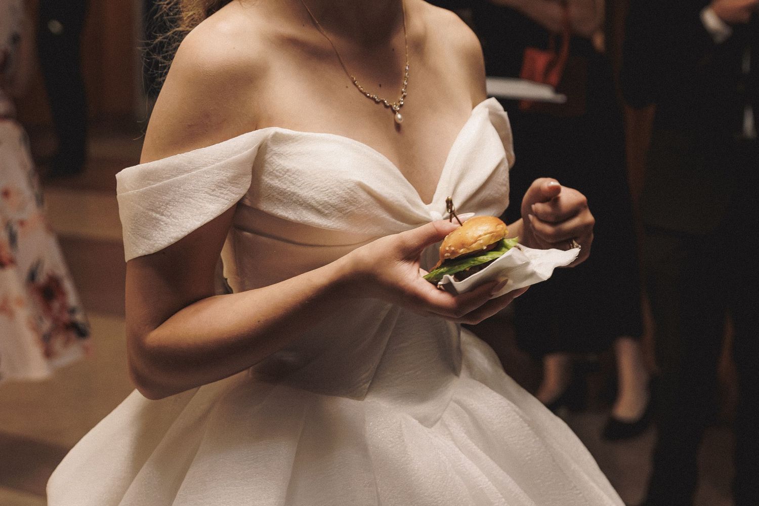 Bride in off-shoulder white tulle gown holds a burger during her wedding reception celebration.