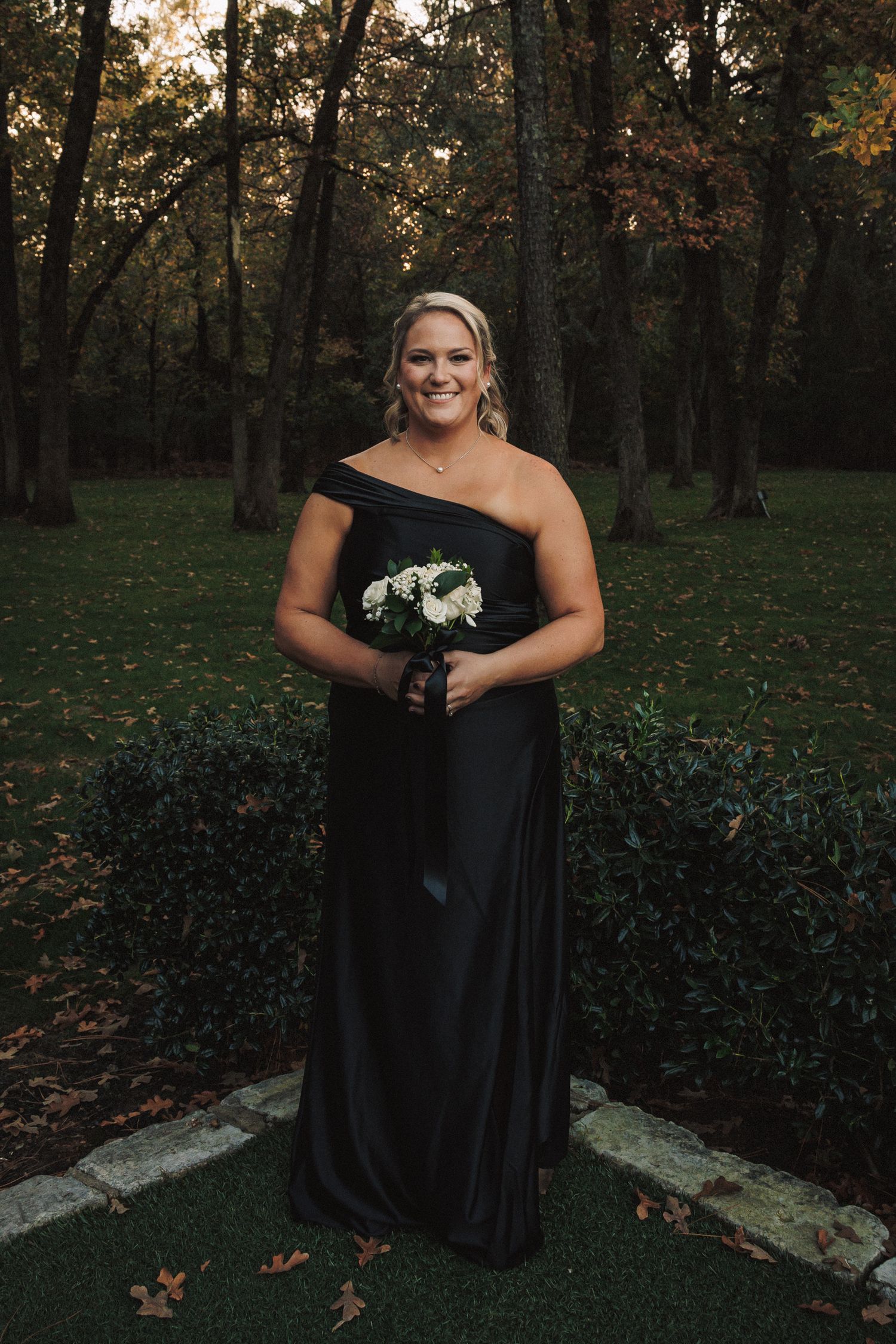 Smiling bridesmaid in black off-shoulder gown holding white bouquet standing in autumn garden.