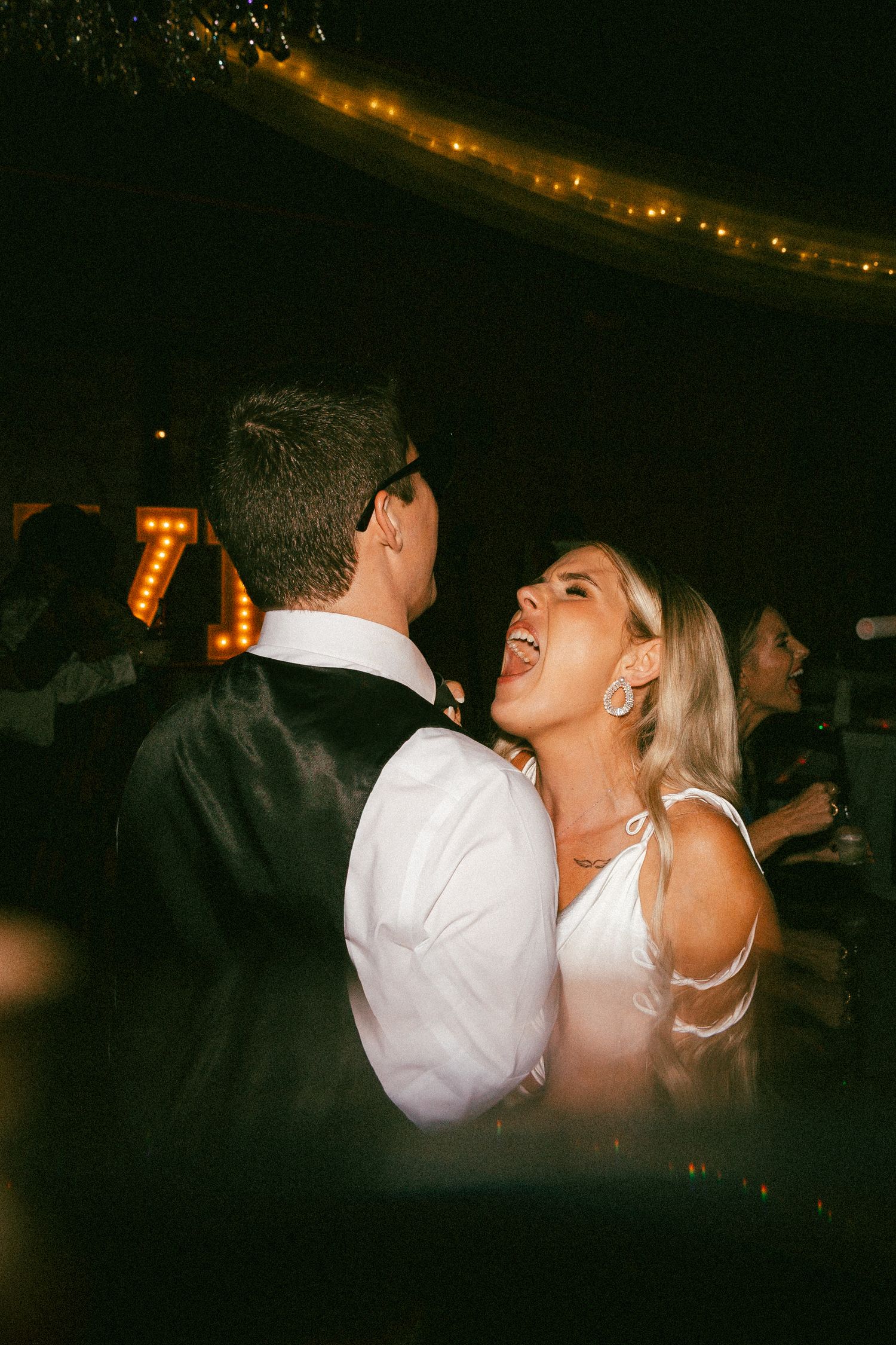 Bride in white gown laughing joyfully while dancing with groom at evening wedding reception.