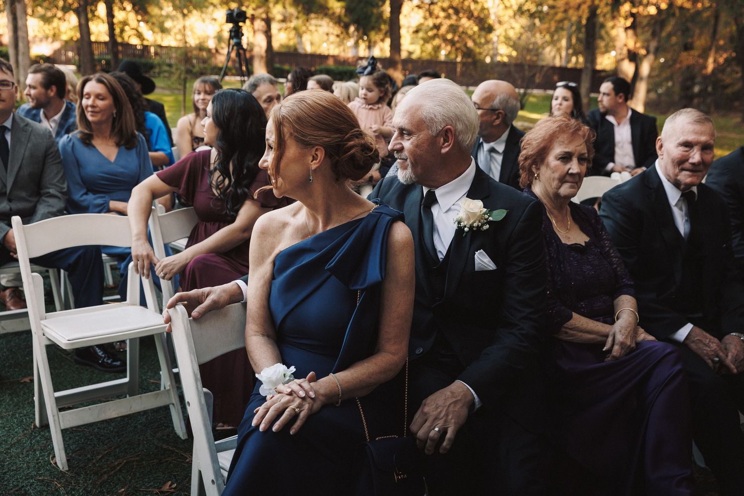 Elegantly dressed wedding guests seated in white chairs during an outdoor autumn ceremony.