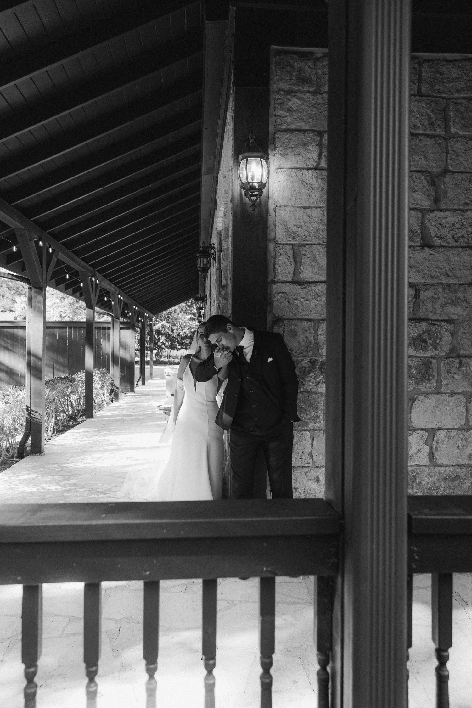 Bride and groom share a romantic kiss on a rustic stone venue porch in black and white.
