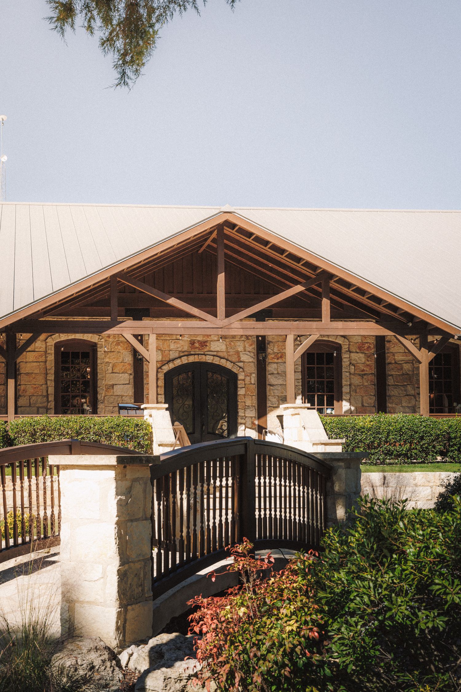 Rustic stone and wood wedding venue exterior with arched entrance, wooden gate, and manicured hedges under clear blue sky.