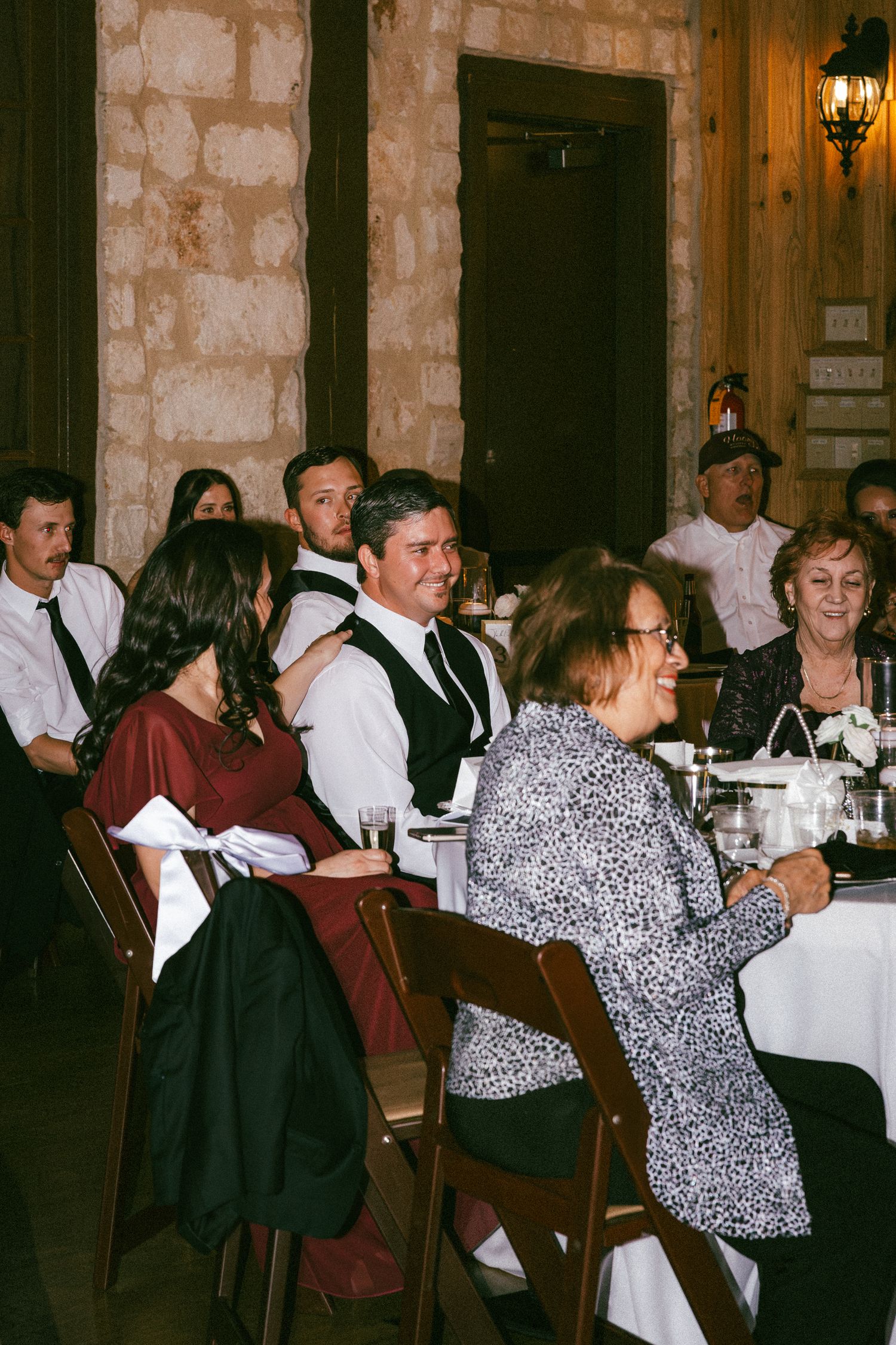 Wedding guests laughing and enjoying dinner reception in elegant stone venue with warm lighting.