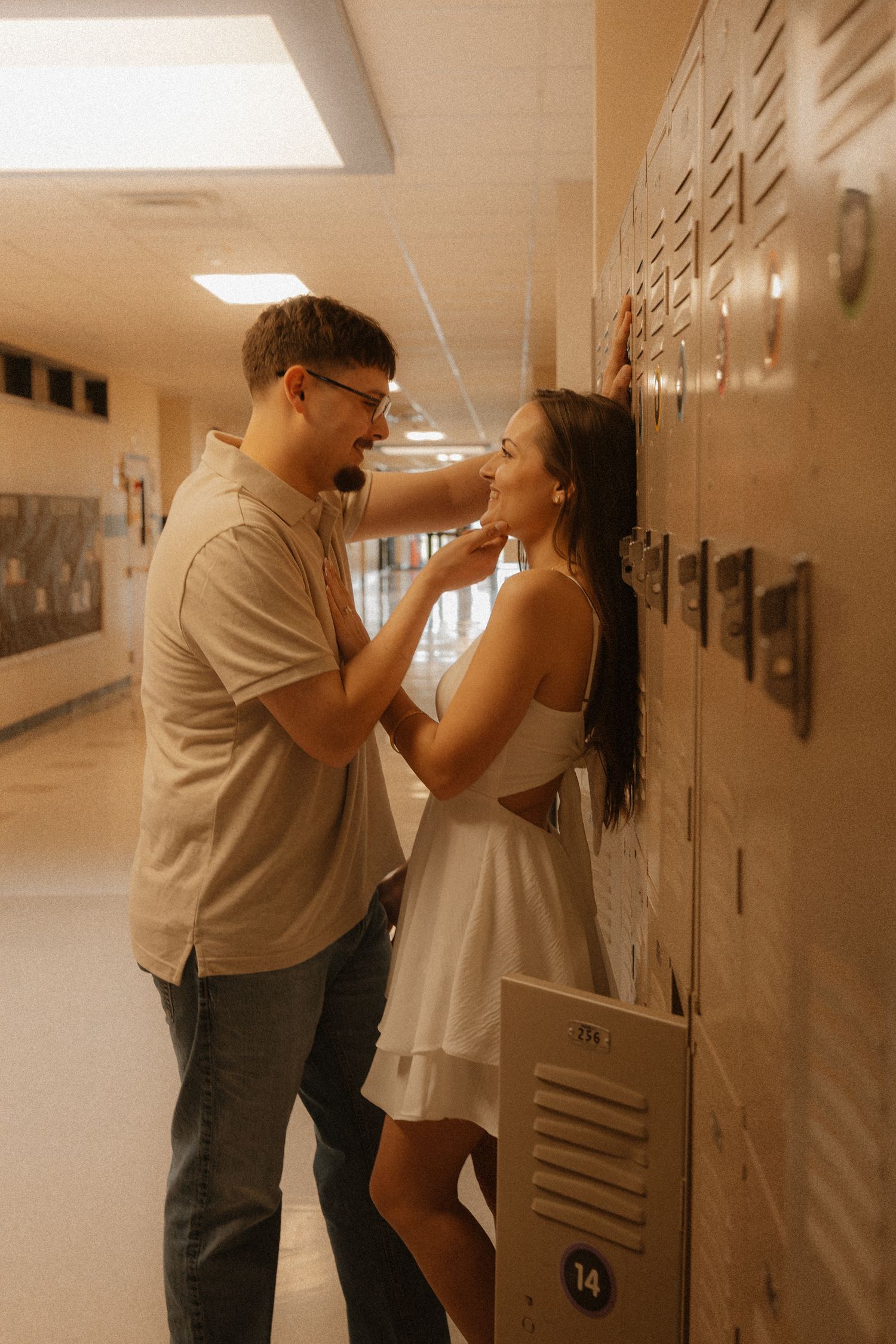 A couple shares a romantic moment in a school hallway, leaning against lockers, with the man gently touching the woman's face.