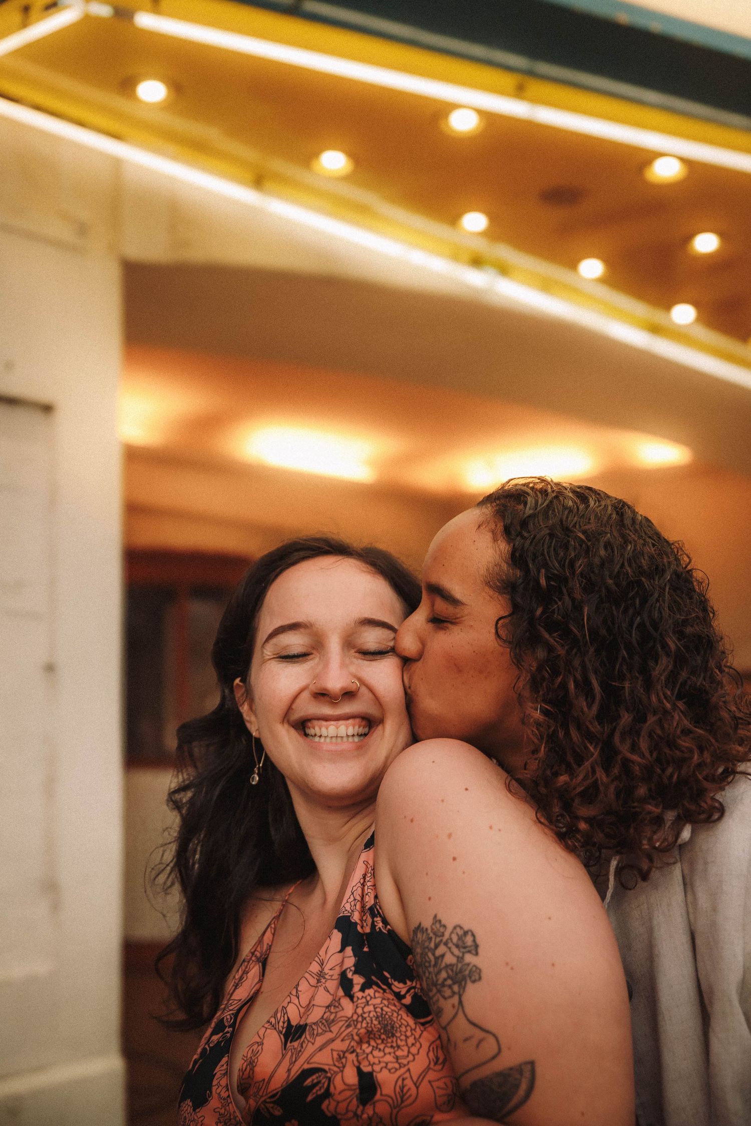 Joyful woman smiles as her partner kisses her cheek under warm neon theater lights after a marriage proposal.
