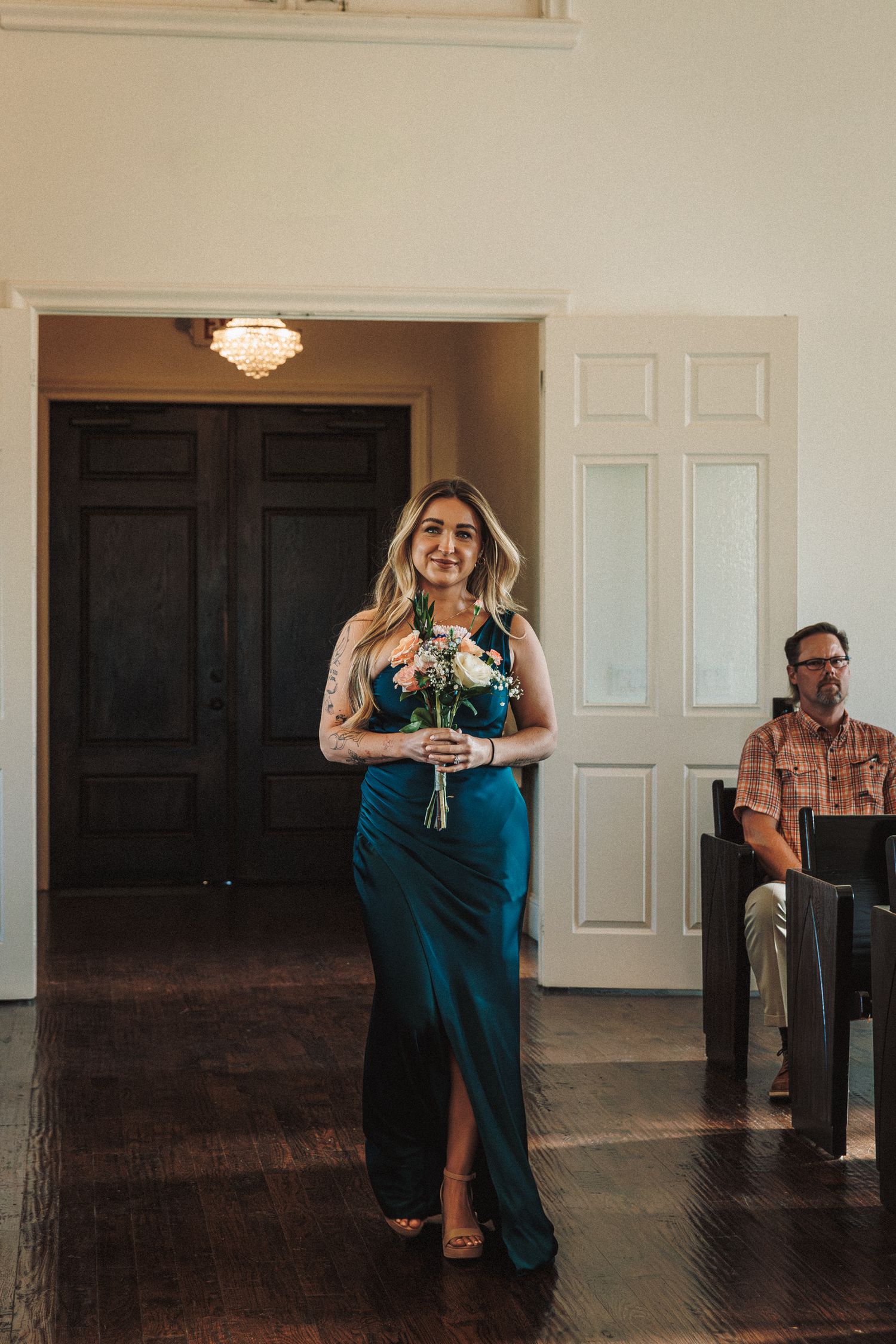 Bridesmaid in teal velvet gown walks down aisle carrying white floral bouquet during wedding ceremony.
