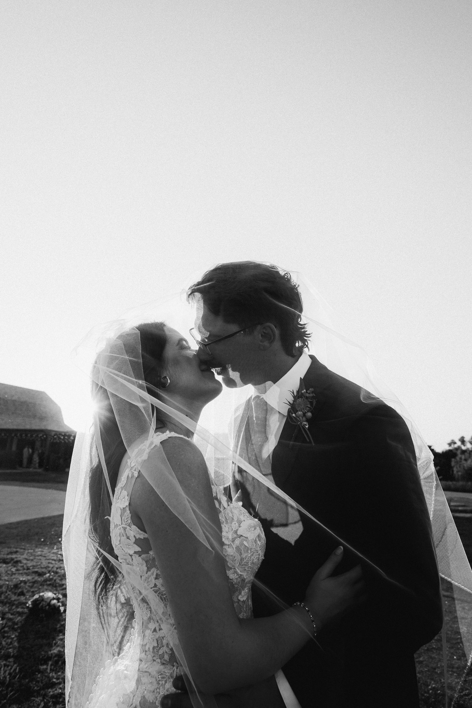 Black and white photo of bride and groom kissing under veil at sunset wedding ceremony.