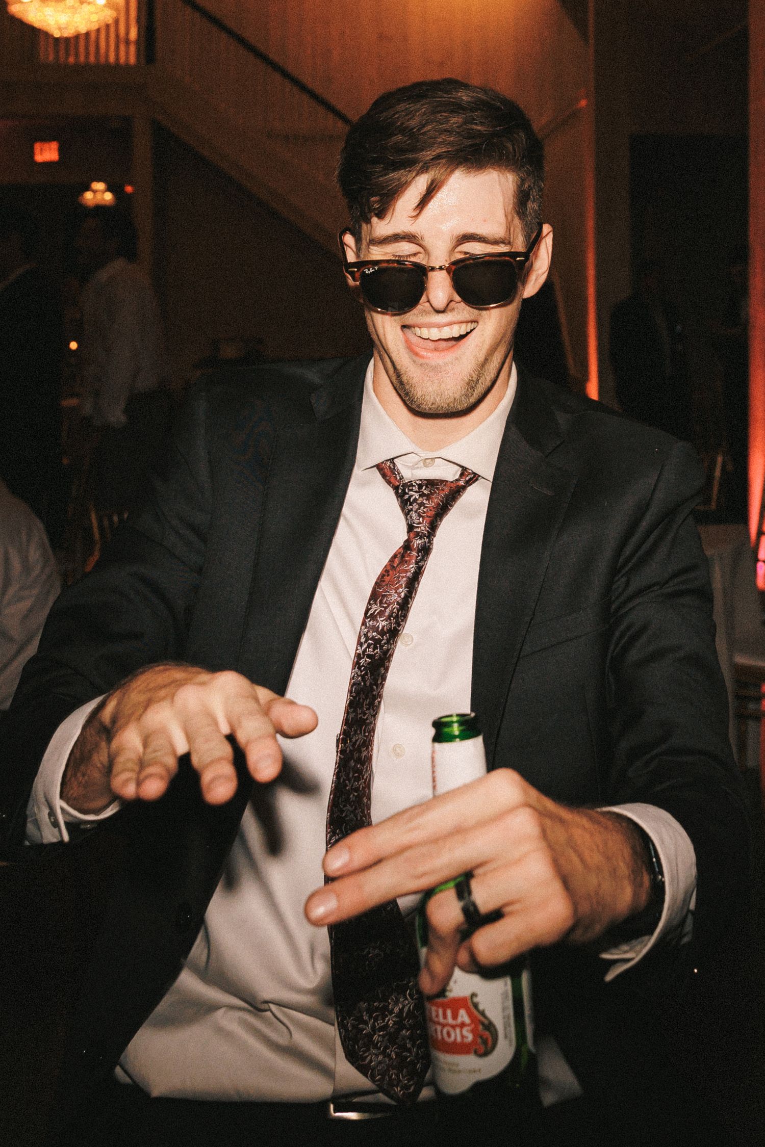 A smiling man in sunglasses and a suit holds a Stella Artois beer bottle at a wedding reception.