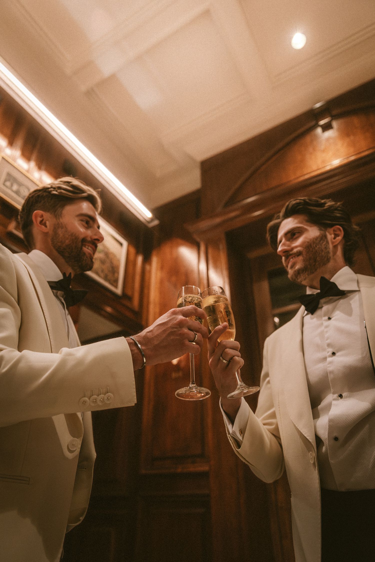 Two grooms in ivory tuxedos toasting champagne glasses in elegant wood-paneled room at wedding reception.