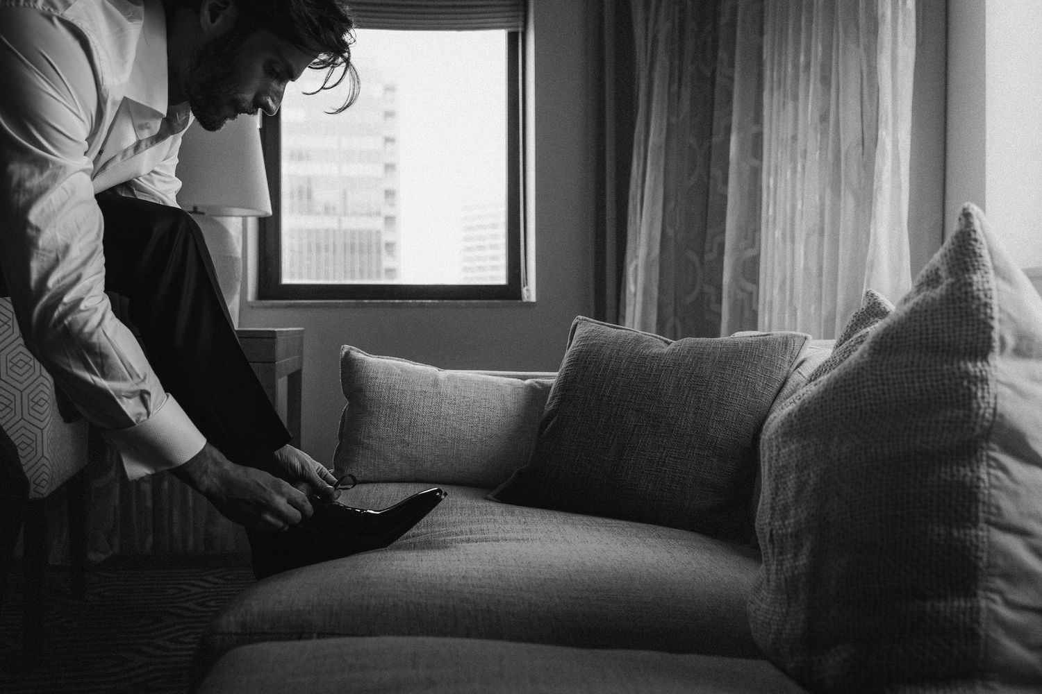 Groom in white dress shirt putting on patent leather shoes beside a gray sofa in a hotel room.