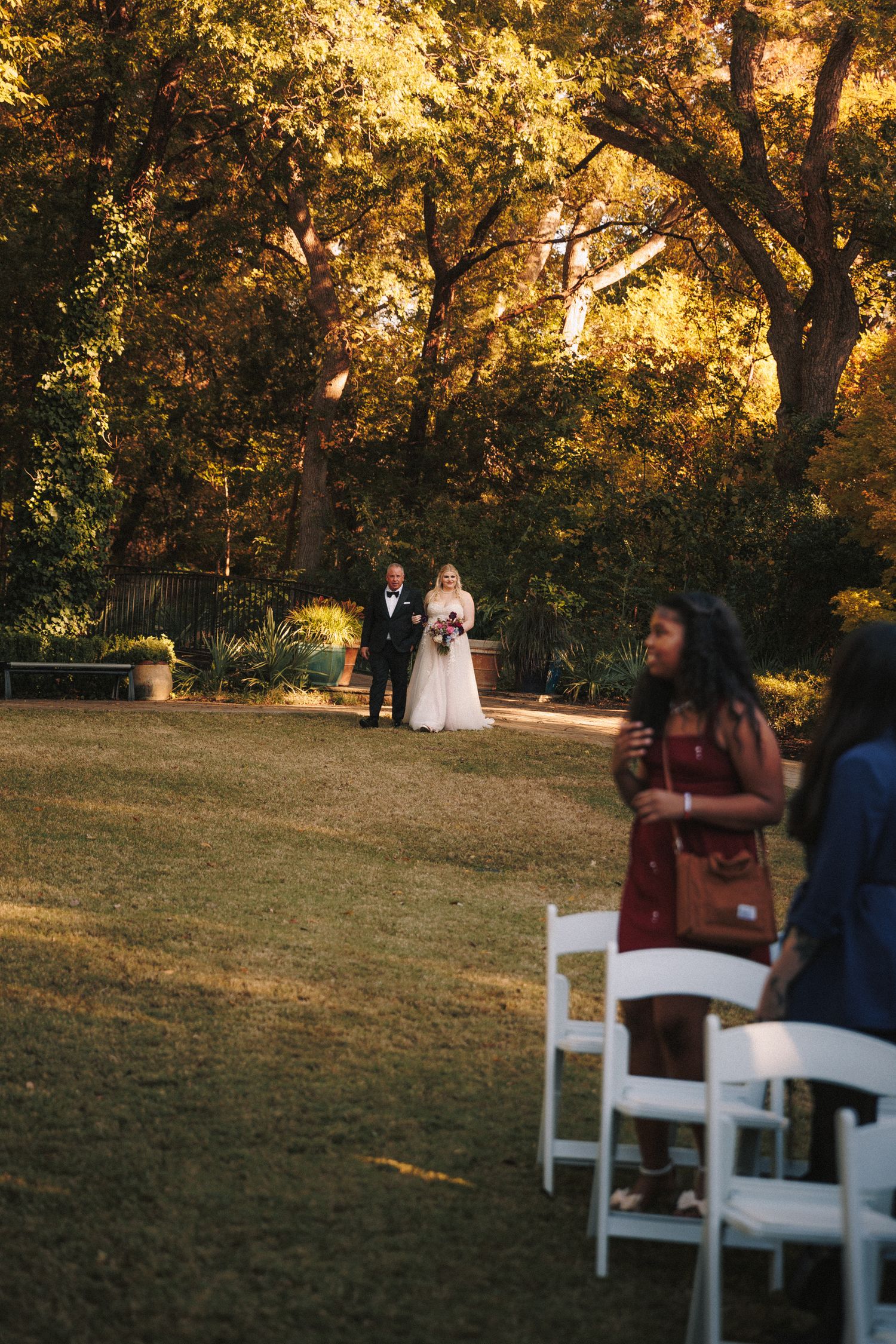 Bride and escort walk down outdoor aisle toward altar as wedding guests watch in golden evening light.