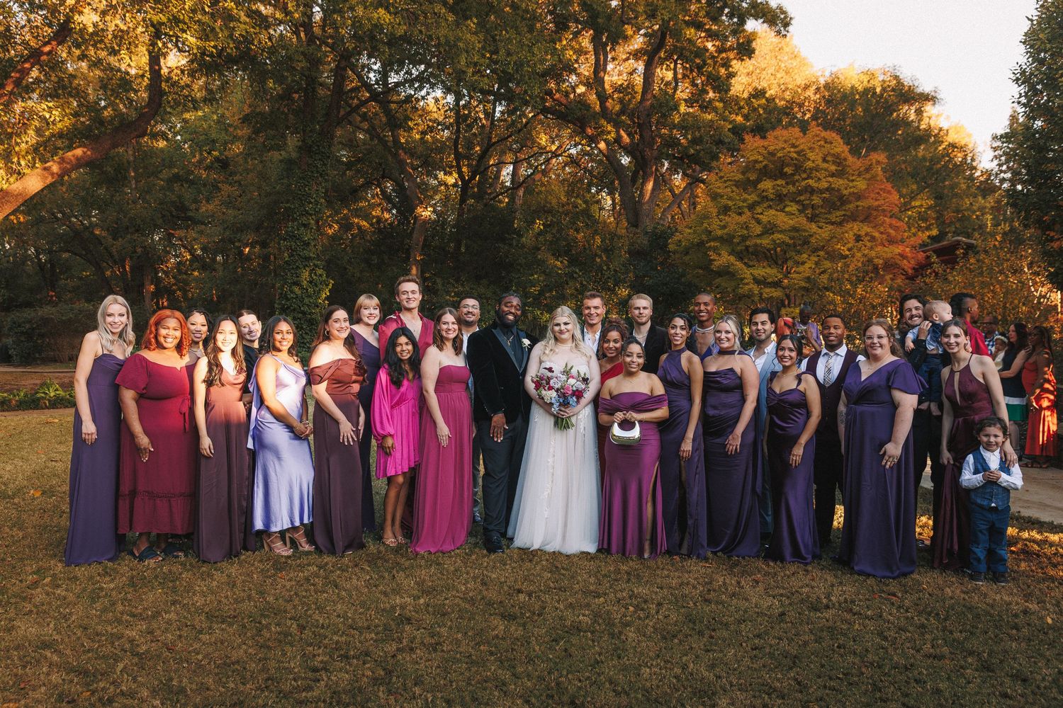 Large wedding party in purple and burgundy gowns posing together in autumn garden setting.
