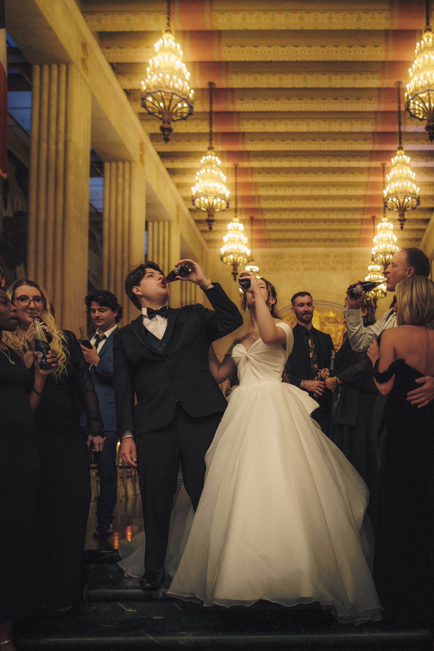Bride and groom celebrating with champagne under ornate chandeliers surrounded by wedding guests.
