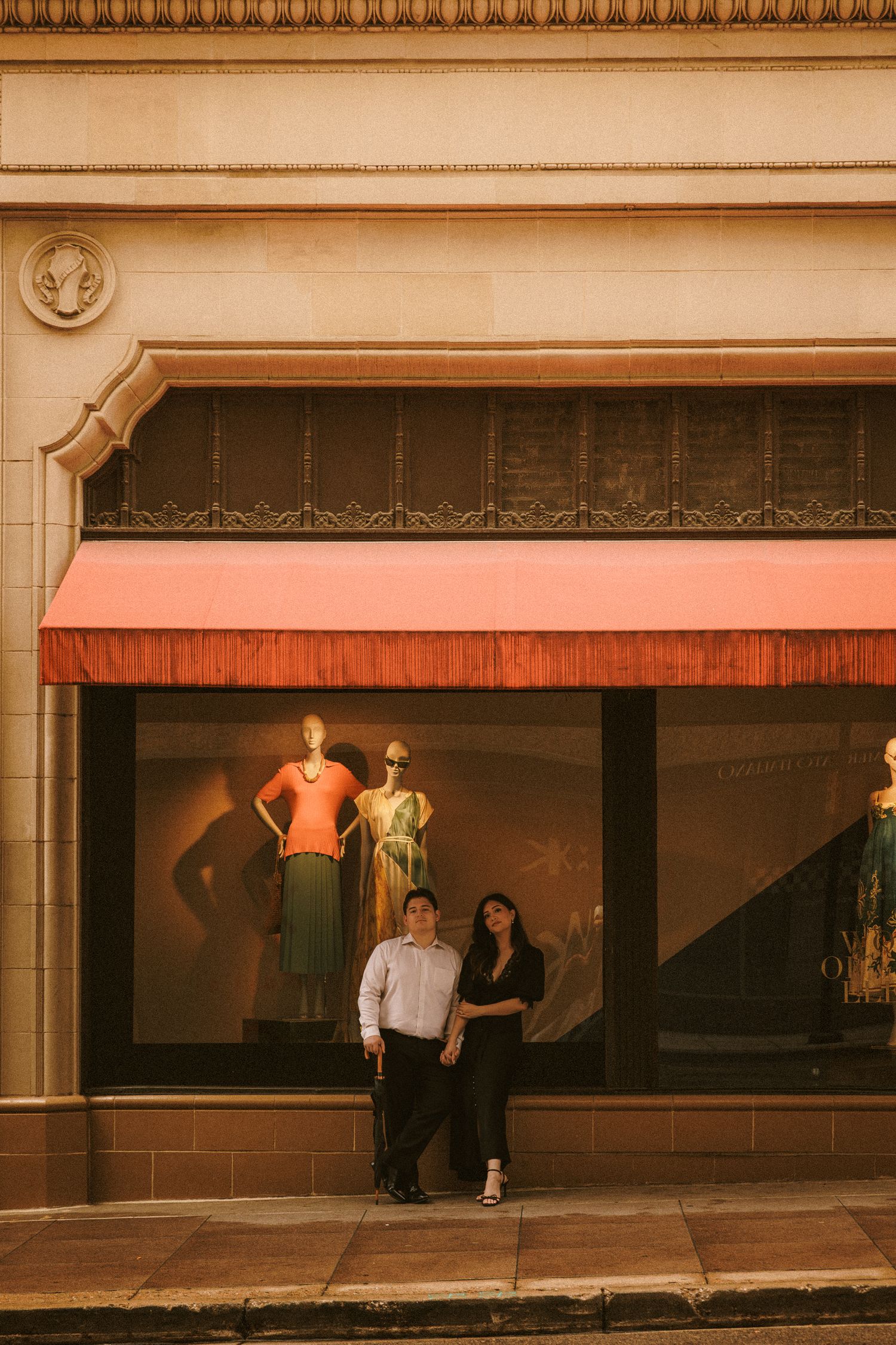An engaged couple poses together in front of an elegant boutique storefront with a red awning.