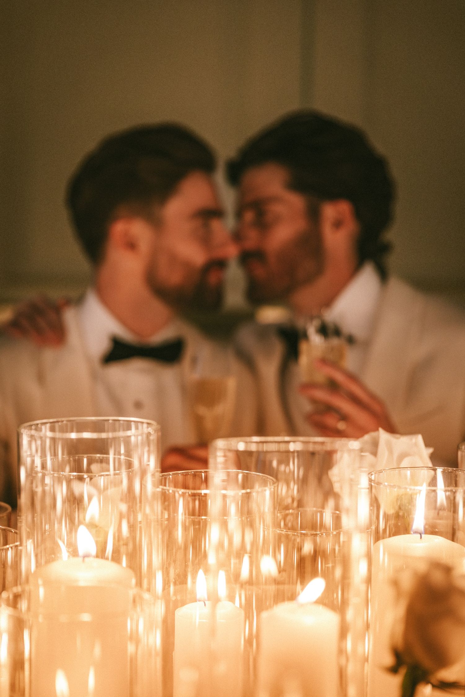 Same-sex couple nearly kissing at candlelit wedding reception table with soft bokeh candle foreground.