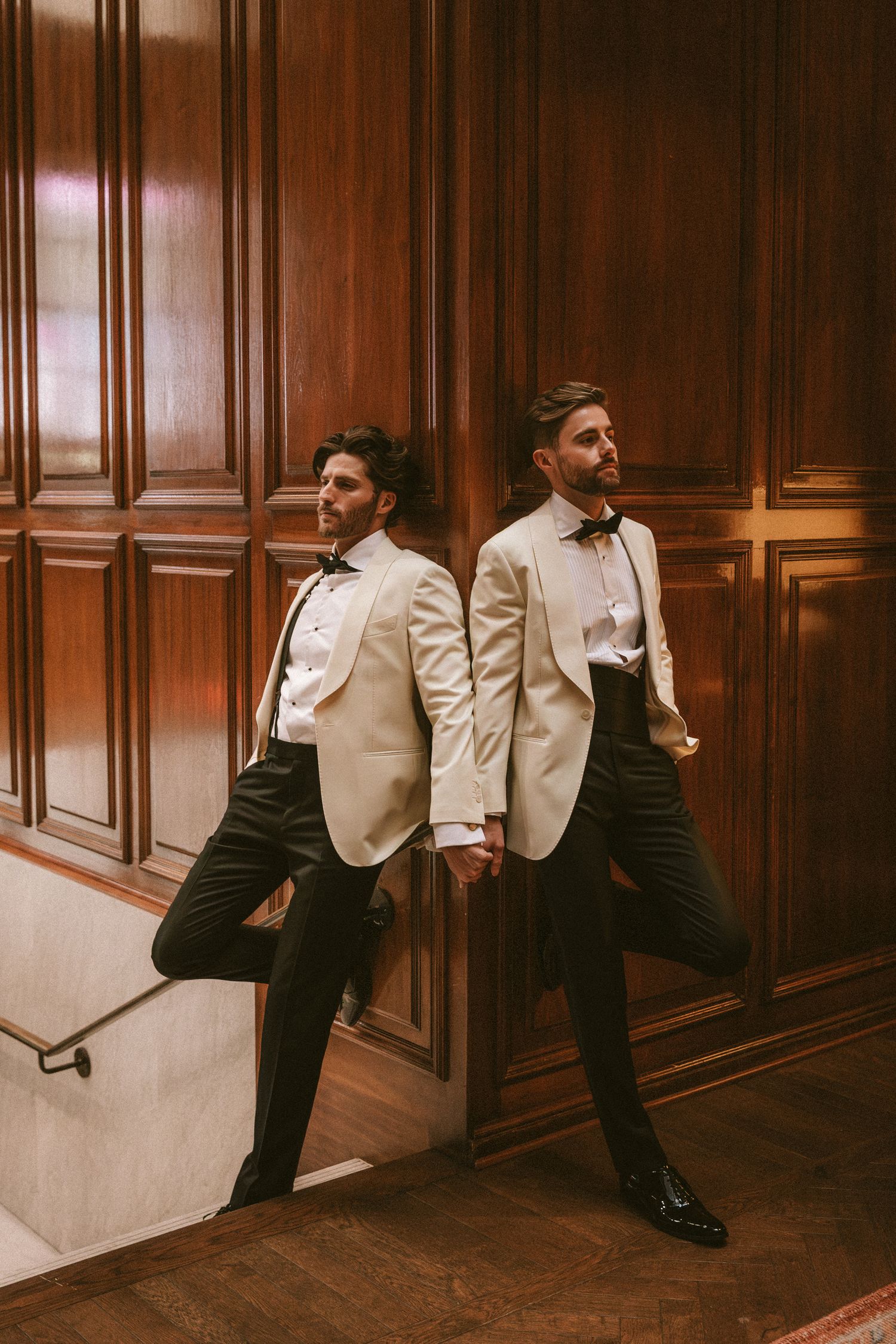Two grooms in white tuxedo jackets lean against rich wood-paneled walls holding hands.
