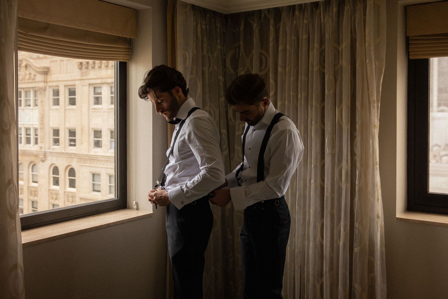 Two groomsmen in white shirts and suspenders helping each other dress.