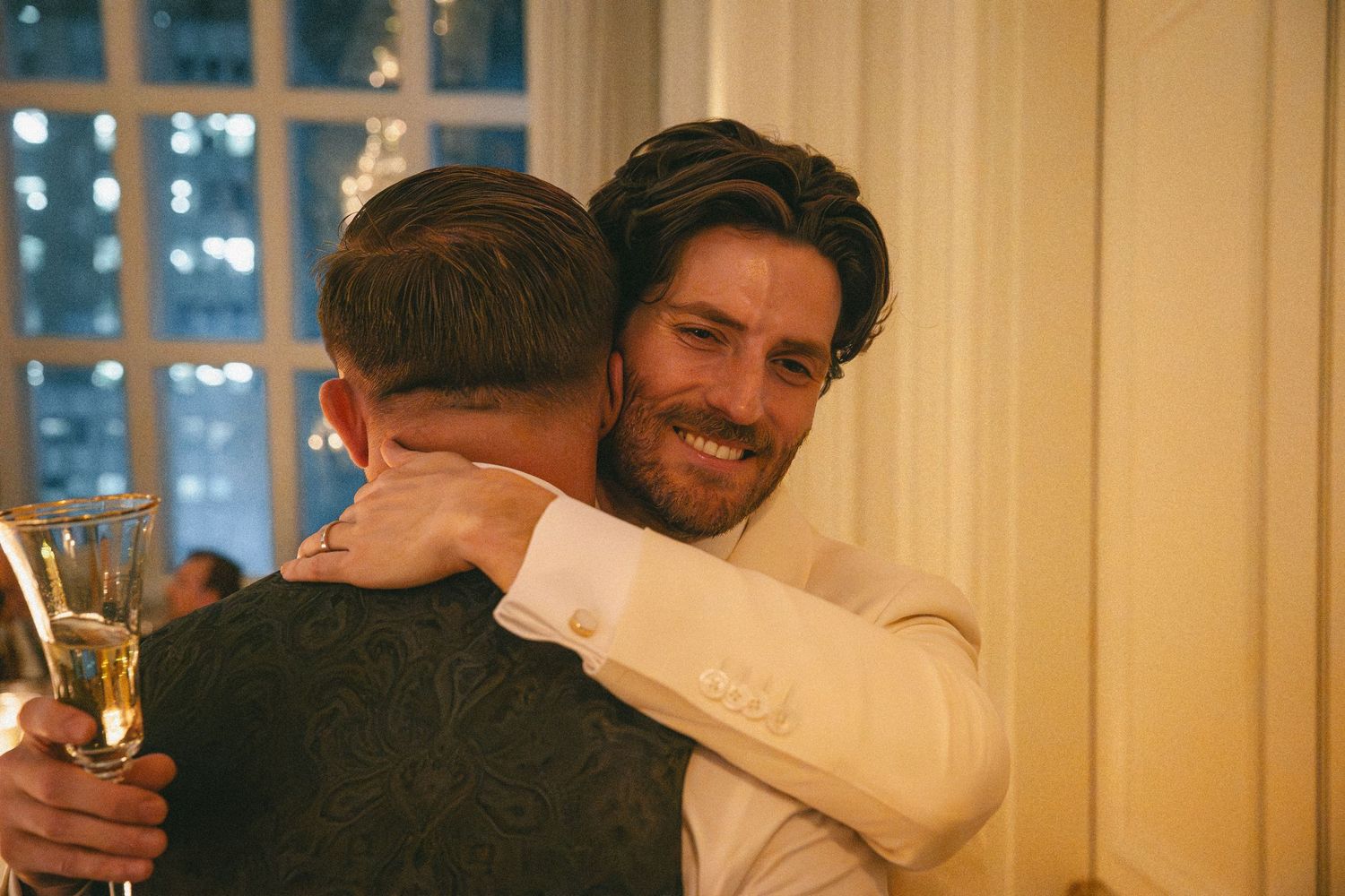 Two men sharing a warm hug at a wedding reception, one holding a champagne glass.