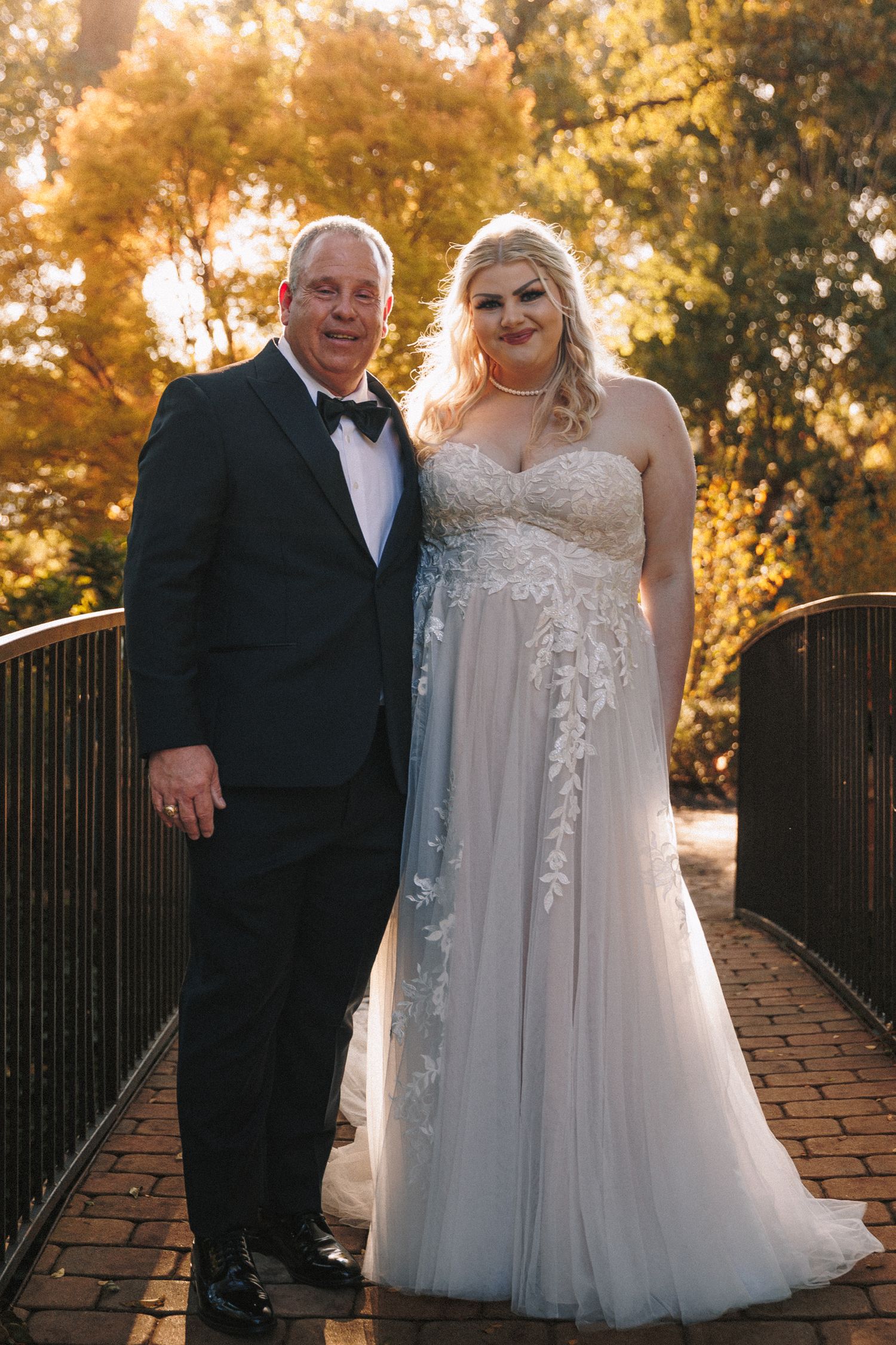Bride in lace wedding gown poses with older gentleman in tuxedo on brick bridge with fall foliage.