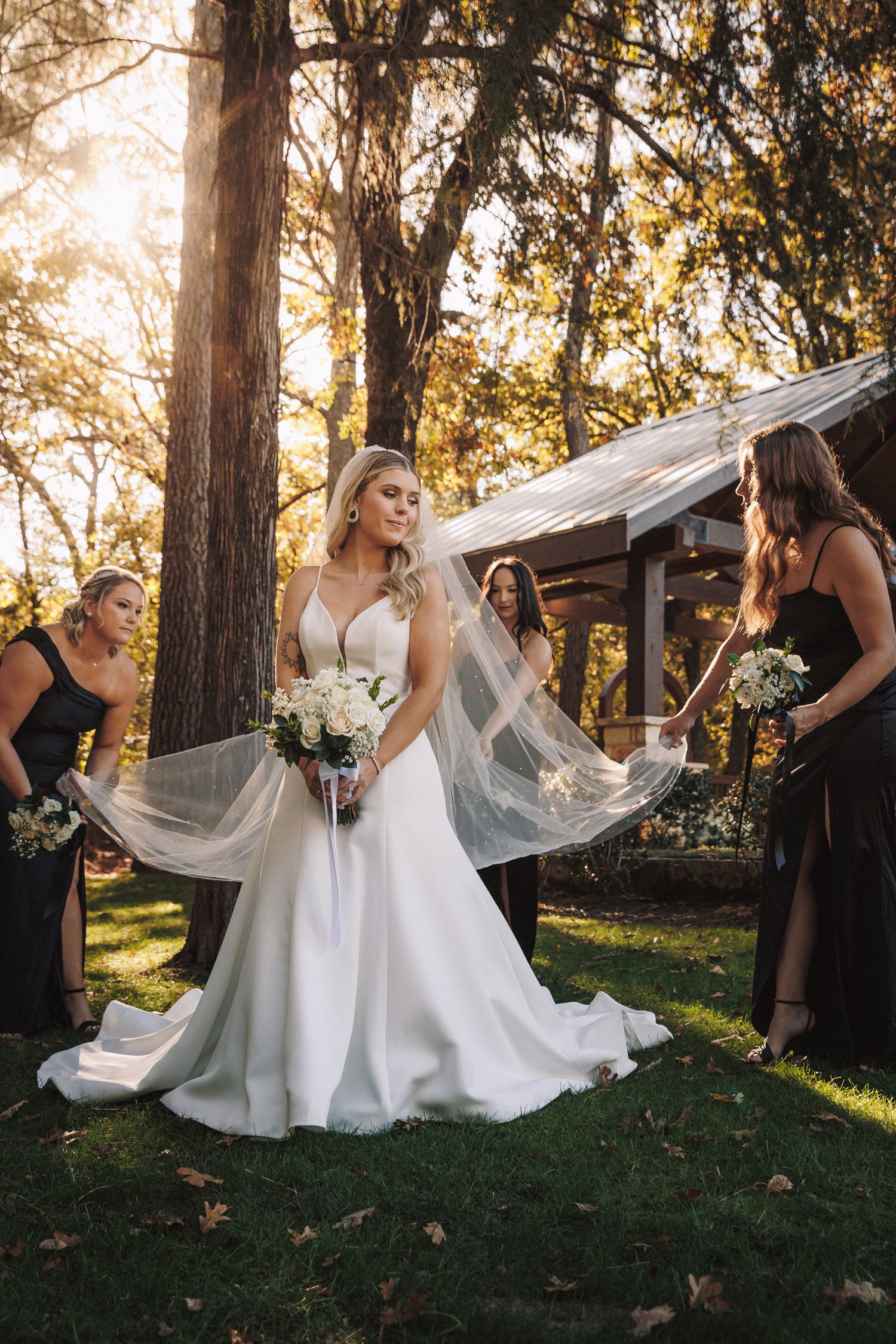 Bridesmaids in black dresses help bride with veil in golden sunset forest wedding setting.