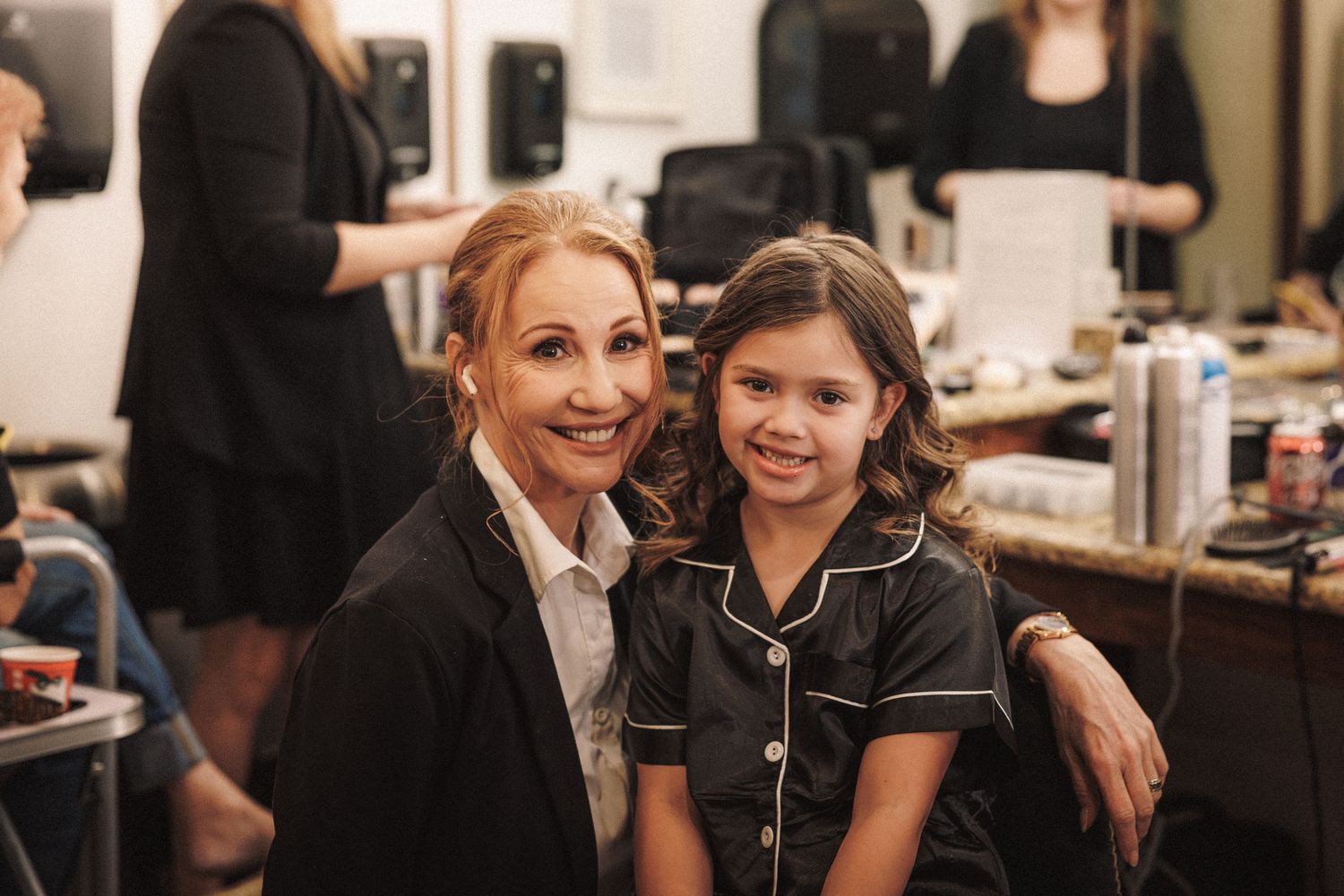 A woman and young girl smile together in matching black outfits in a bridal getting-ready room.