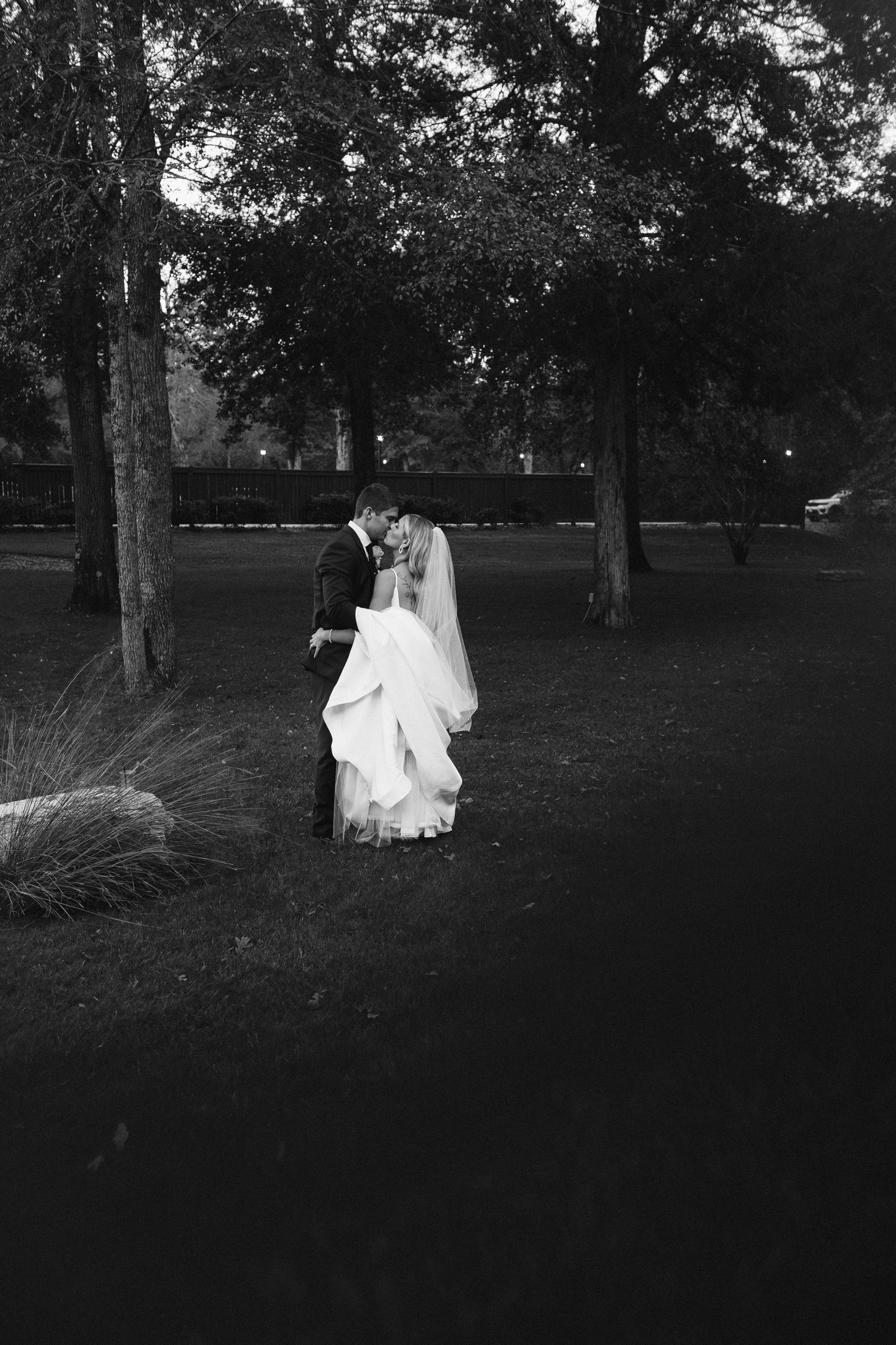 Black and white photo of a bride and groom embracing under trees in a park at twilight.