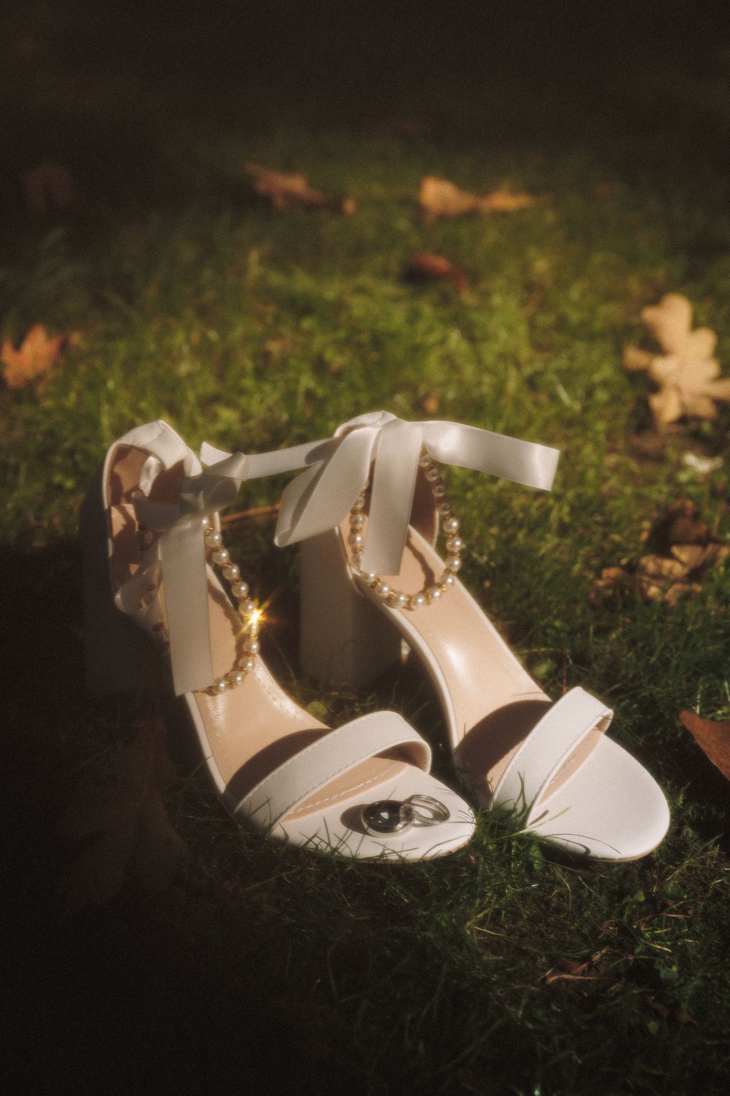 White bridal heeled sandals with pearl ankle straps and satin bow, placed on green grass with autumn leaves.