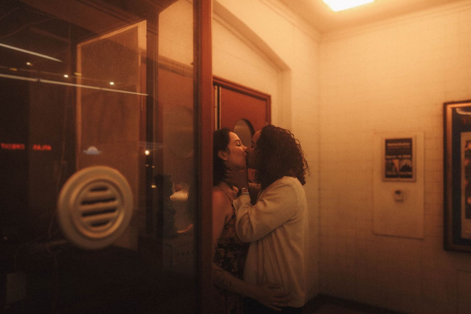Two people sharing an intimate moment near a glass door in a warmly lit indoor space at night.