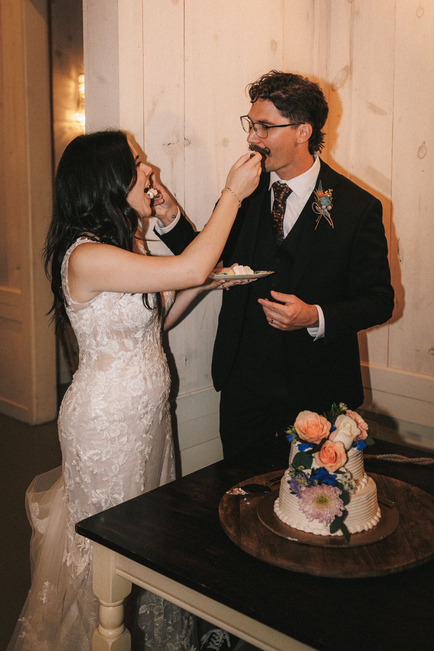 Bride feeds groom a slice of wedding cake decorated with peach and blue flowers at reception.