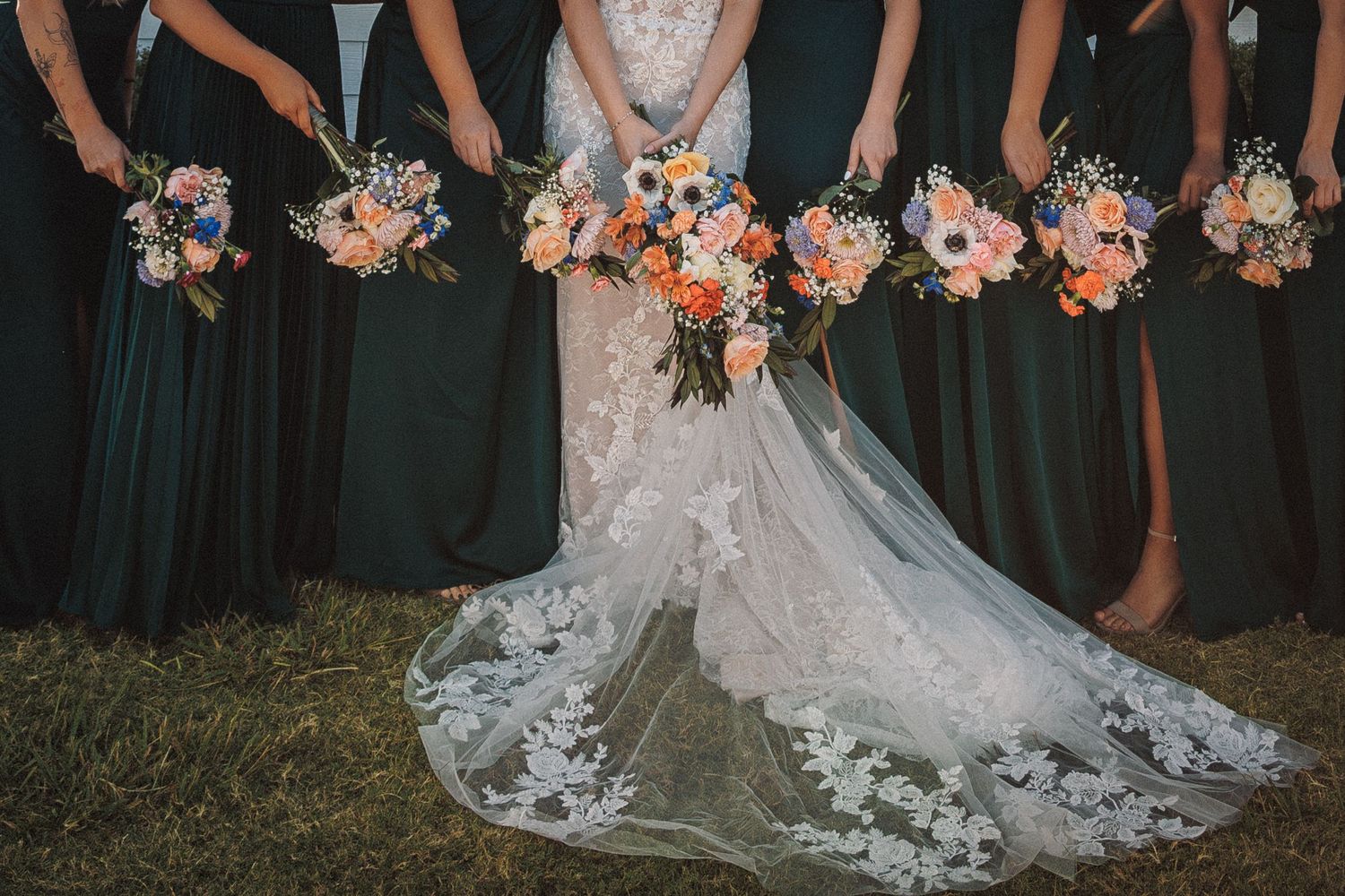 Close-up of bride in lace gown and bridesmaids in emerald dresses holding colorful peach and pink bouquets.