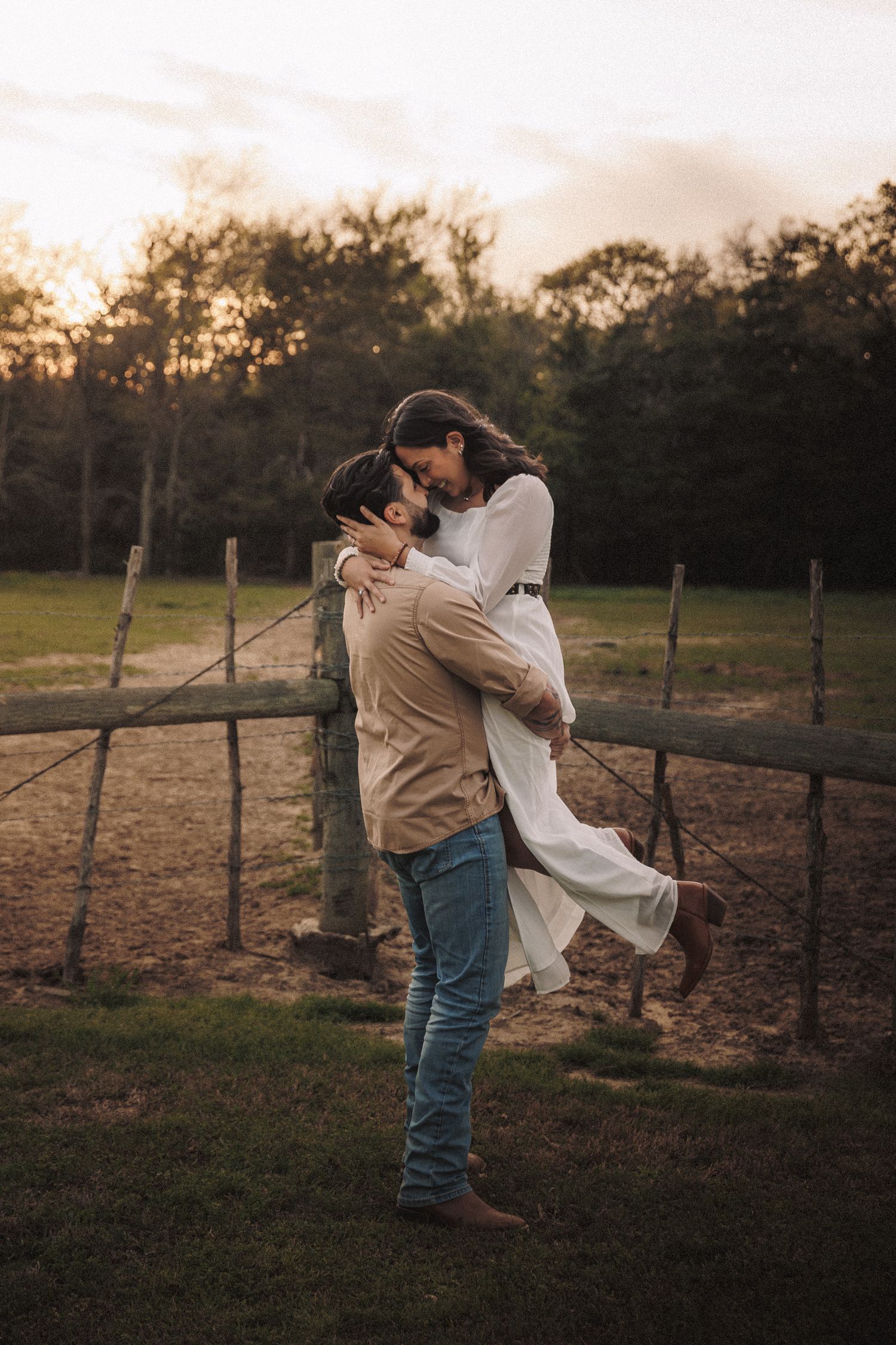 Couple sharing romantic embrace at sunset near rustic wooden fence in green field.