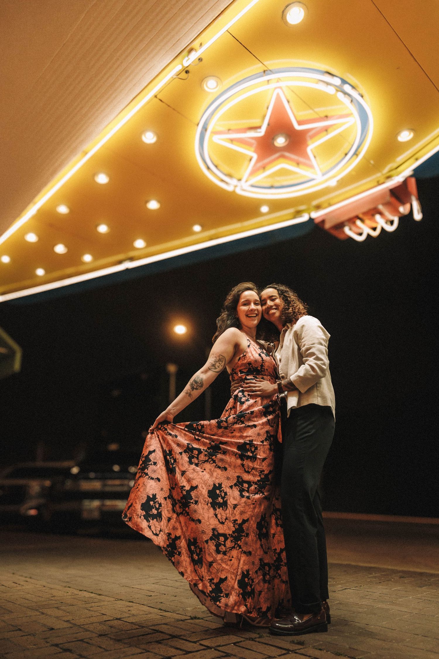 Couple embraces and dances under vibrant neon star sign outside a theater at night, woman in floral dress.