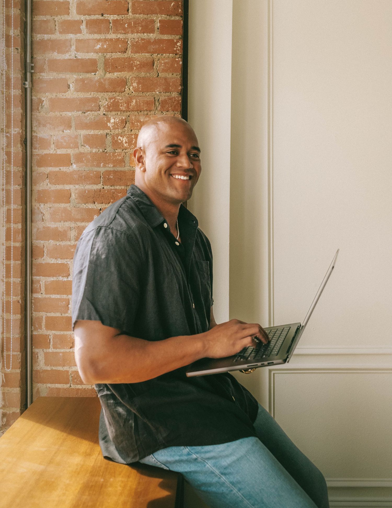 Person in a black shirt works on laptop while sitting comfortably on floor against exposed brick wall.