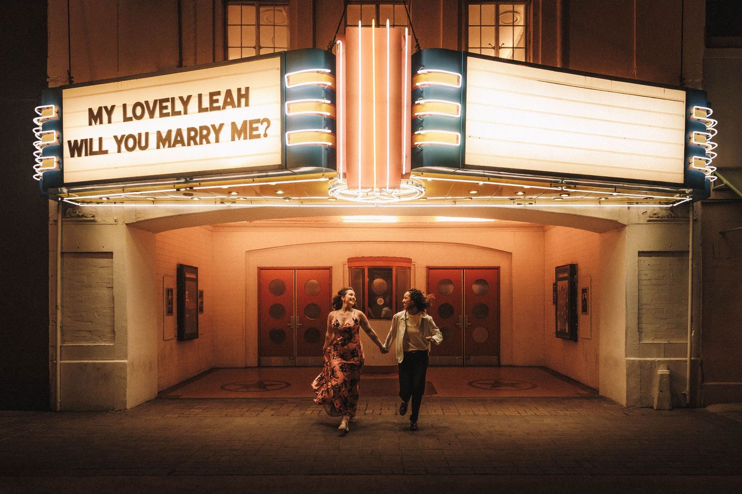 A movie theater marquee displays 'MY LOVELY LEAH WILL YOU MARRY ME?' as a couple stands beneath the romantic proposal.