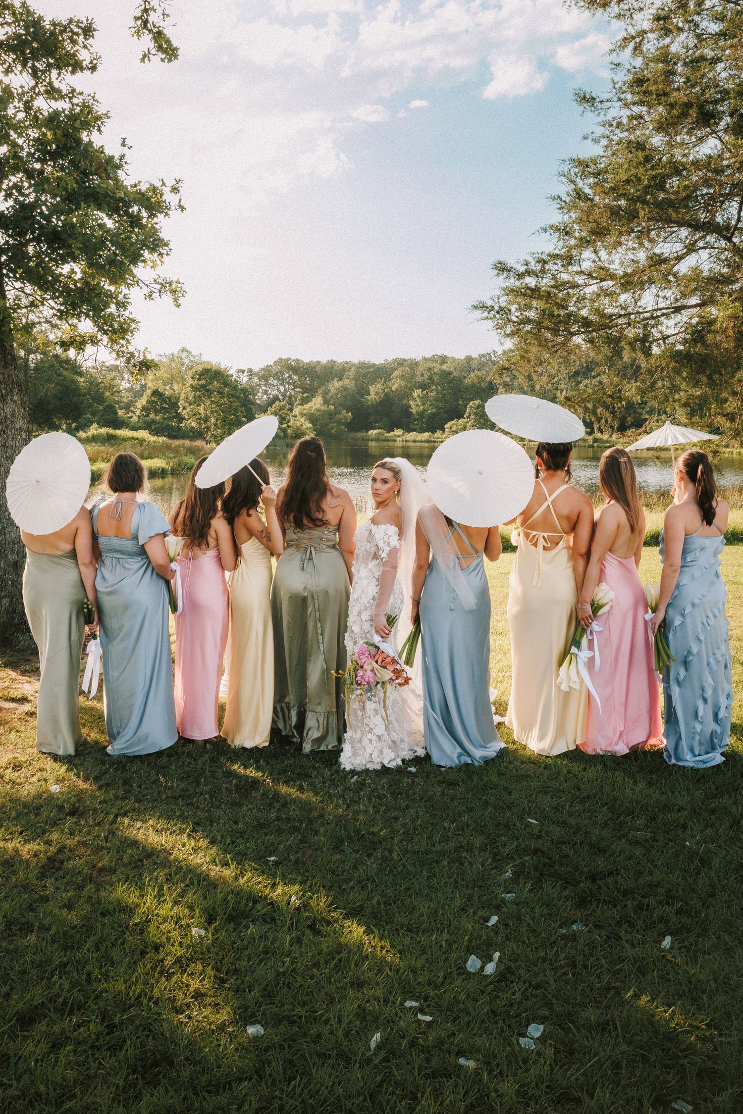 Wedding party lined up in pastel dresses creates whimsical photo on grassy hillside.
