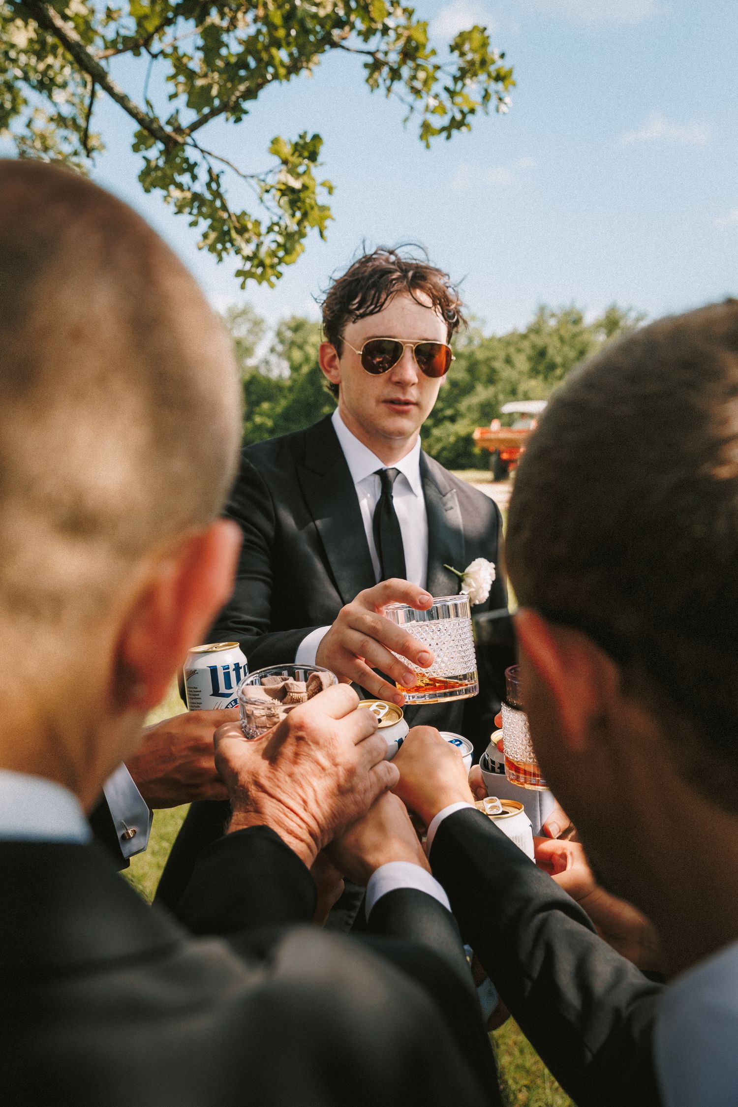 Groomsmen in black suits and sunglasses lighting cigars together outdoors.