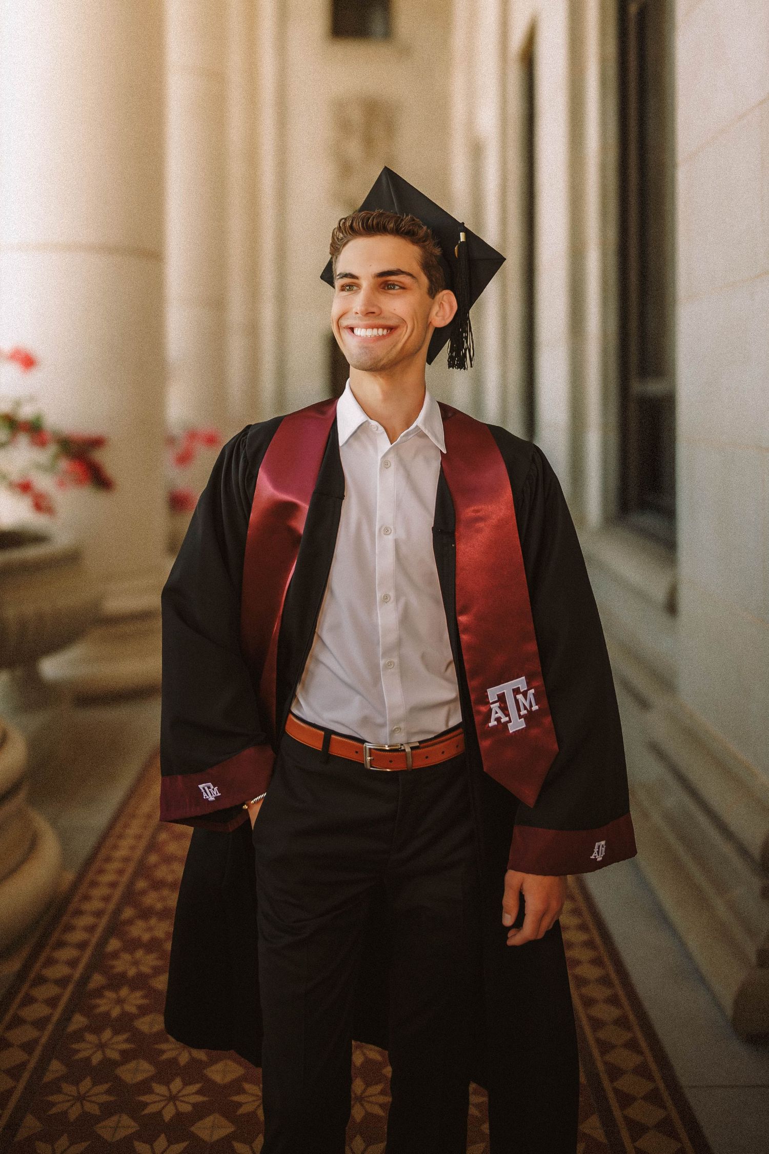 A person in a maroon vest and white shirt walks and smiles down a dimly lit hallway with ornate carpet.