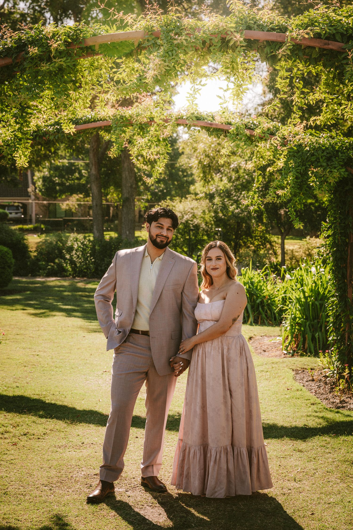 A couple poses together in formal attire in a sunlit garden with lush greenery.