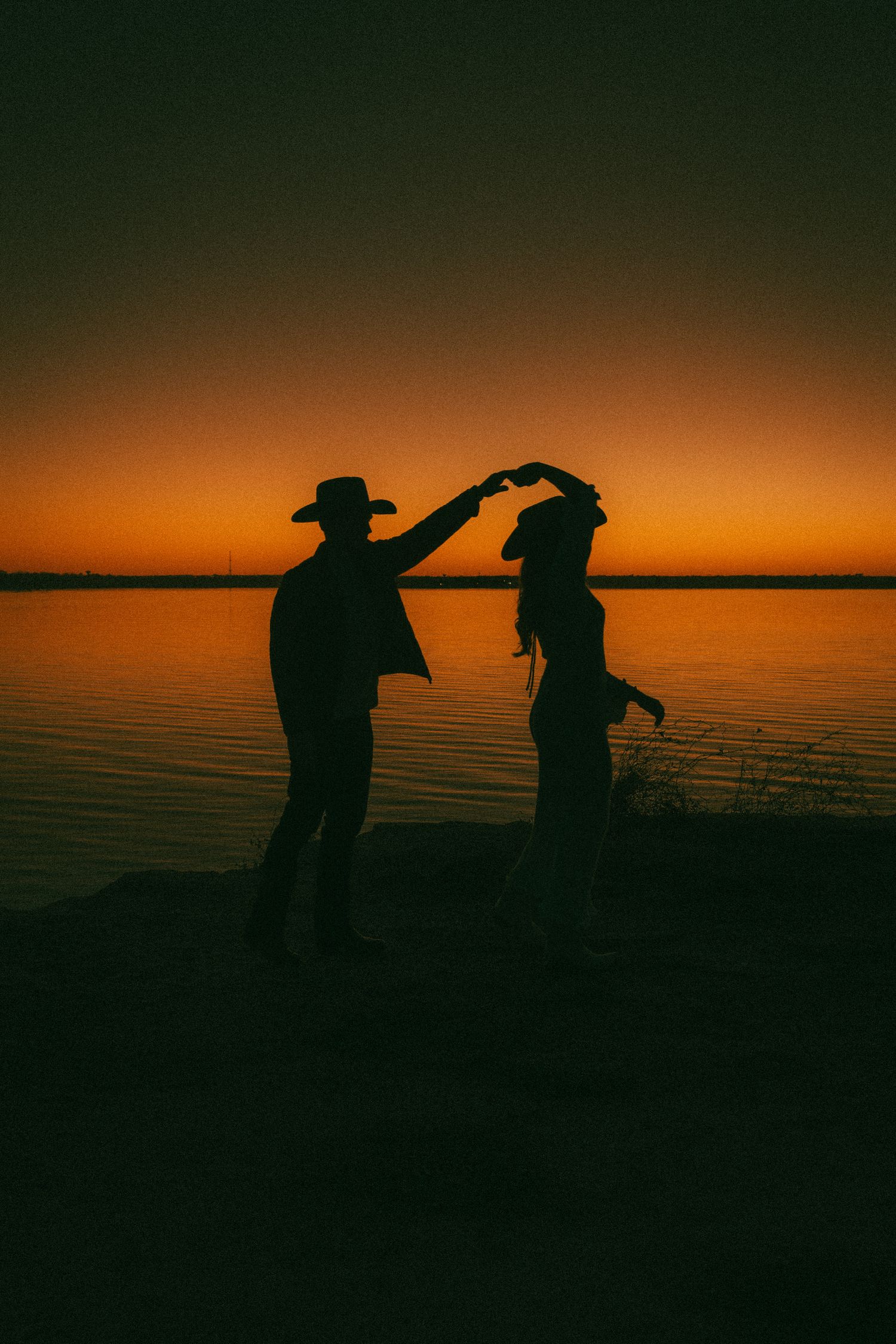 Silhouettes of two people dancing against a dramatic orange sunset over calm waters at dusk.