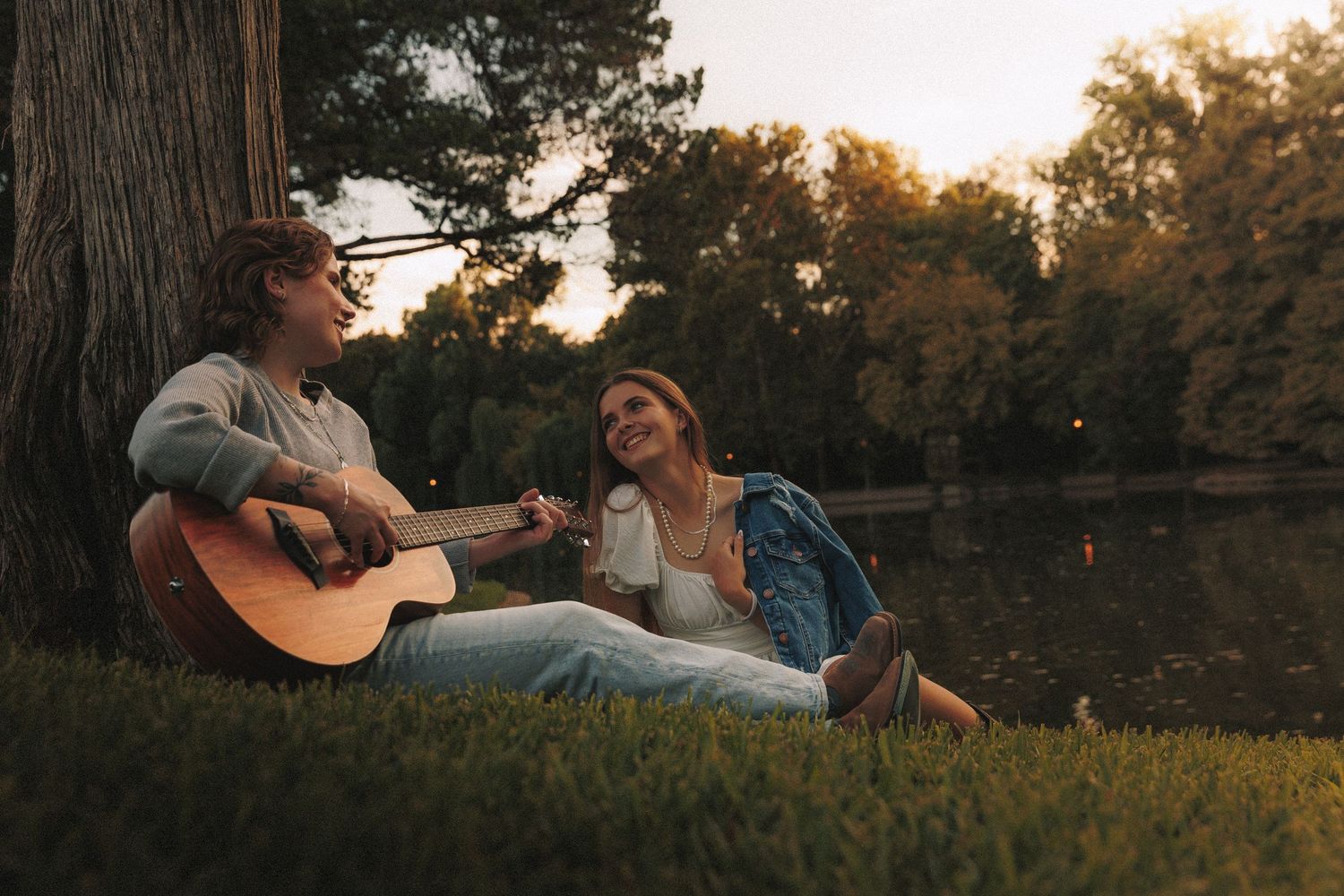 Two people relax by a lake at sunset as one plays acoustic guitar while sitting under a large tree.